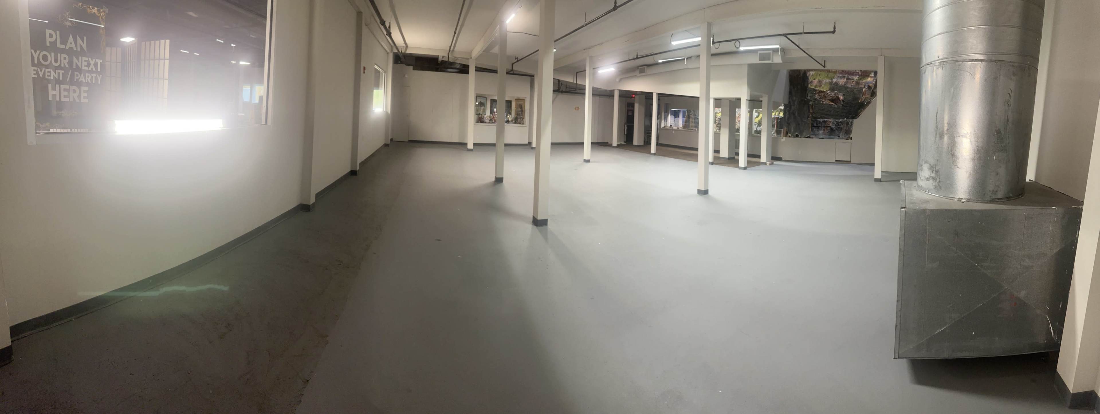 The image shows a large, empty room with gray flooring, white walls, and several support poles throughout the space.