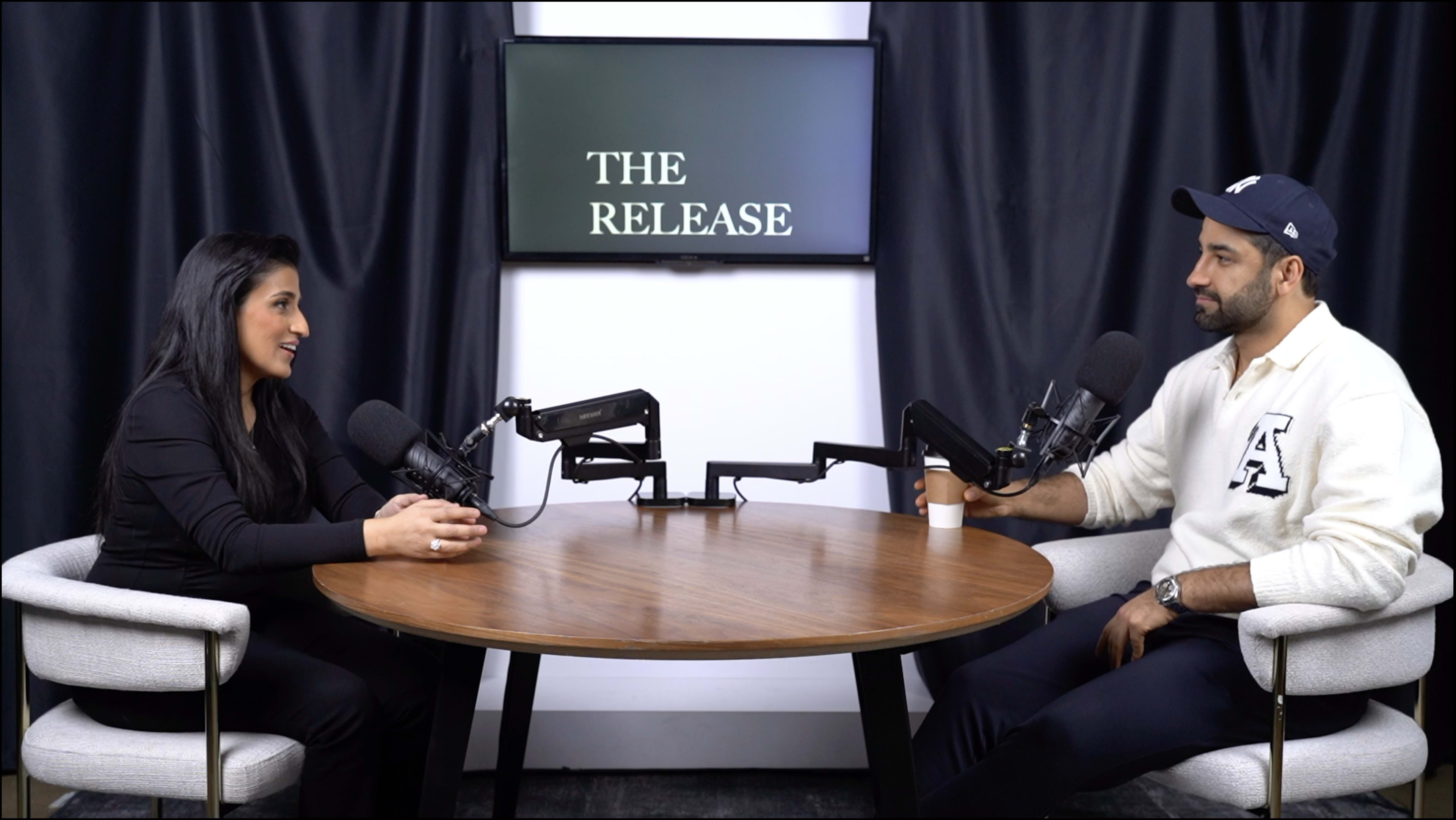 Two individuals are seated across from each other at a round table, engaged in conversation, with microphones positioned in front of them and a display screen behind displaying the text "THE RELEASE."