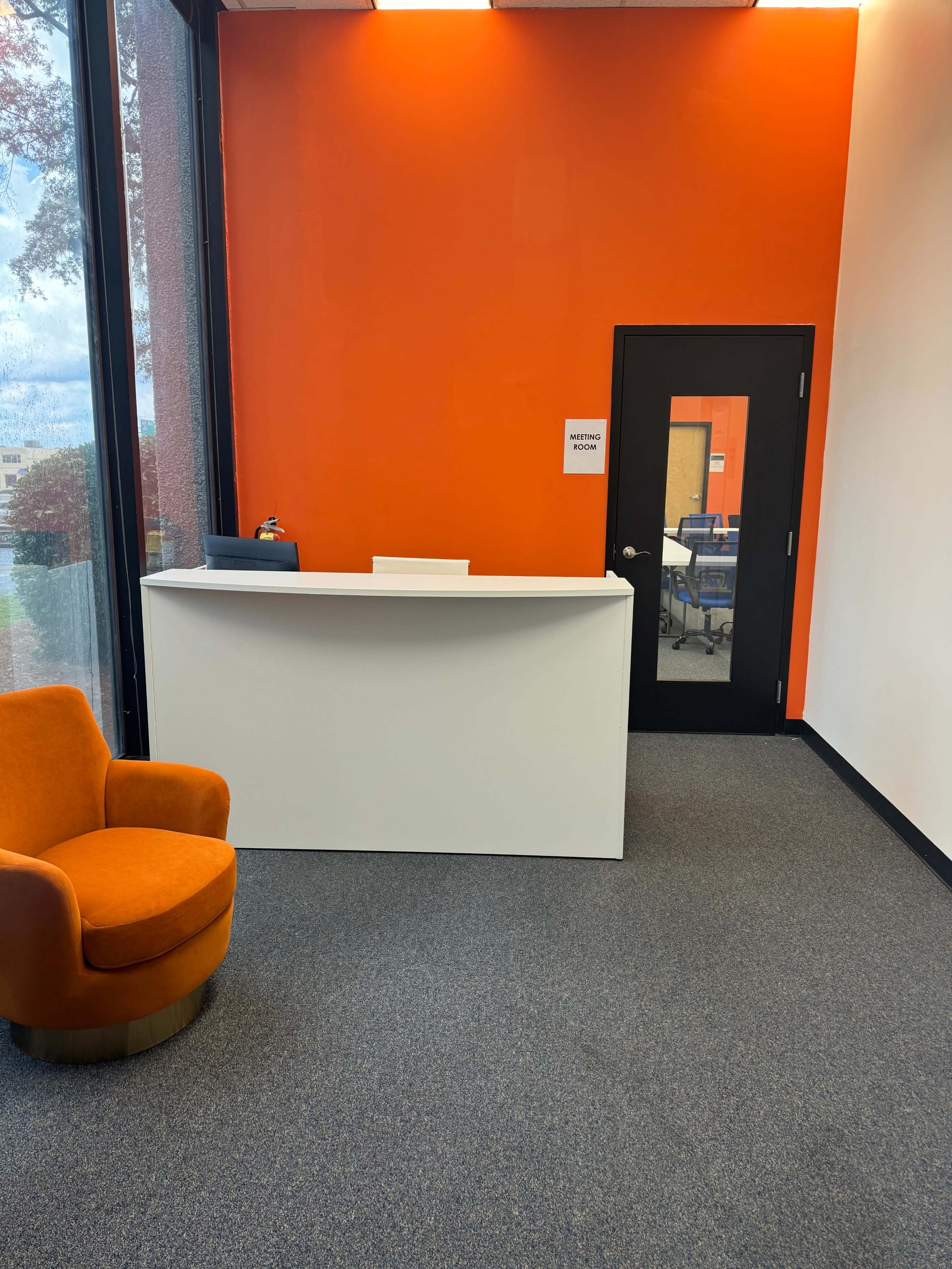 The image shows a modern reception area with an orange accent wall, a white front desk, a black door, and an orange upholstered chair.