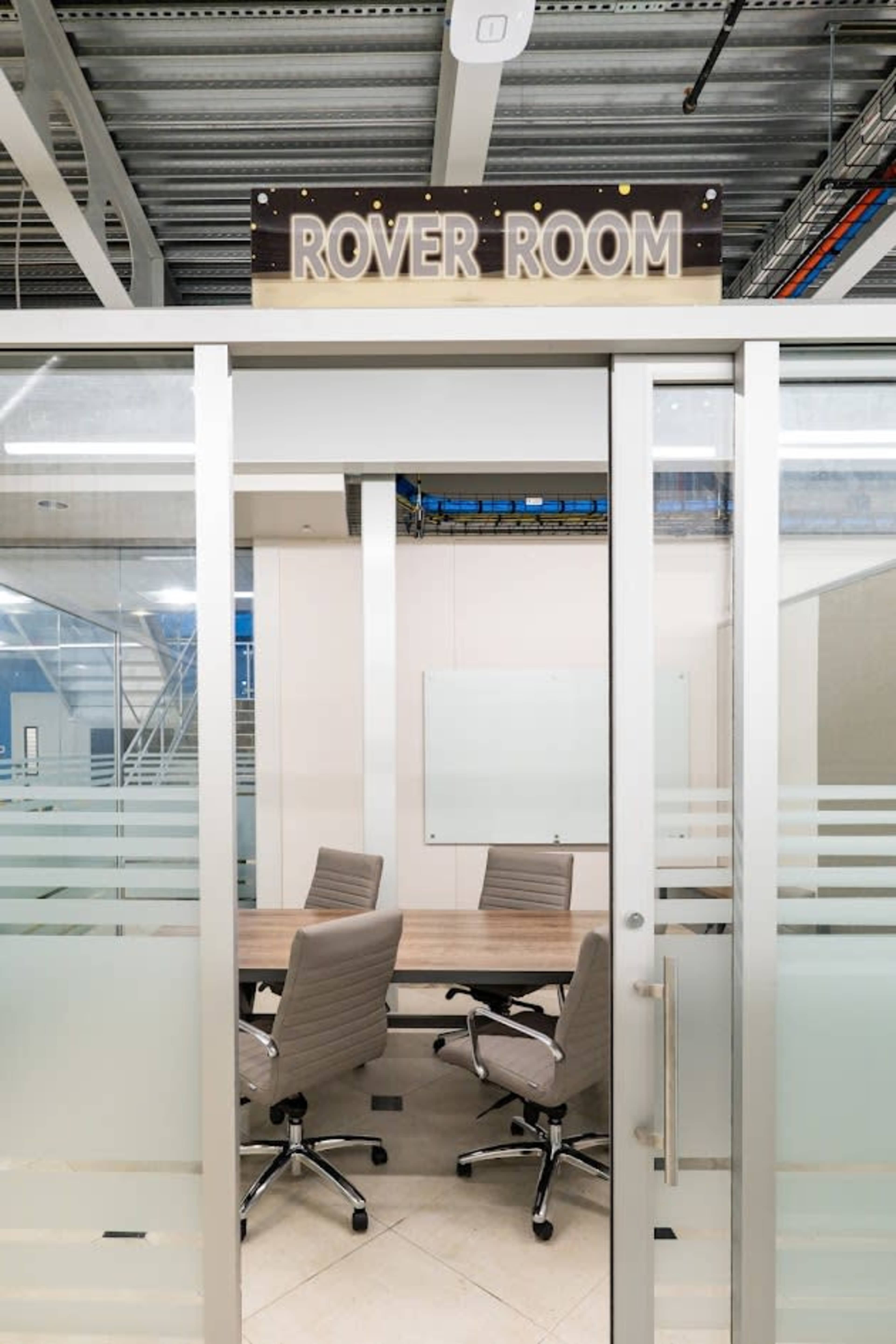The image shows a conference room labeled "ROVER ROOM" with a glass door and four chairs arranged around a table inside.