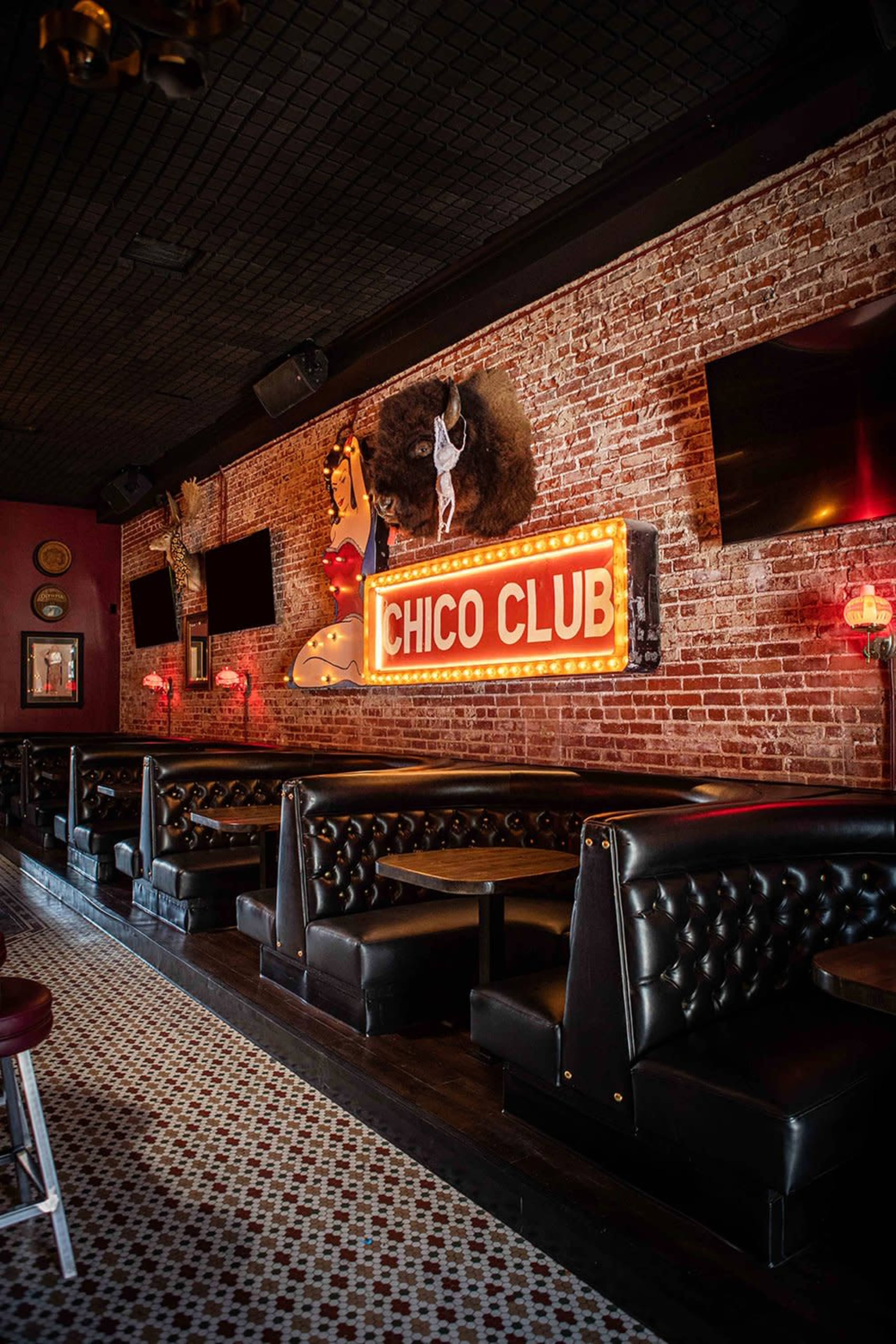 The interior of a bar features a vintage brick wall, black leather seating booths, and a brightly lit neon sign reading "Chico Club."
