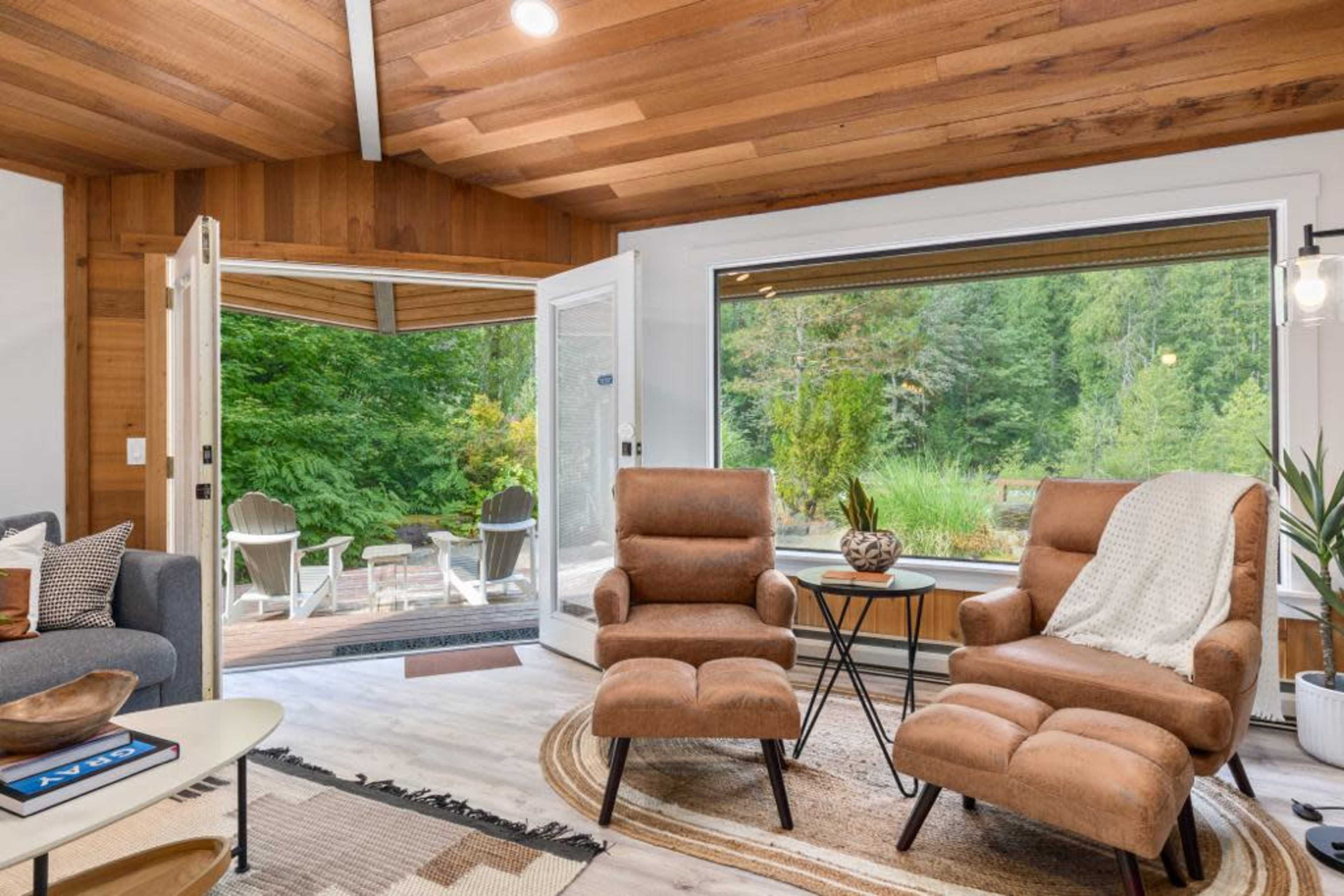 A cozy interior space featuring two brown armchairs, a small side table, and large windows that open to a scenic outdoor view of trees and greenery.