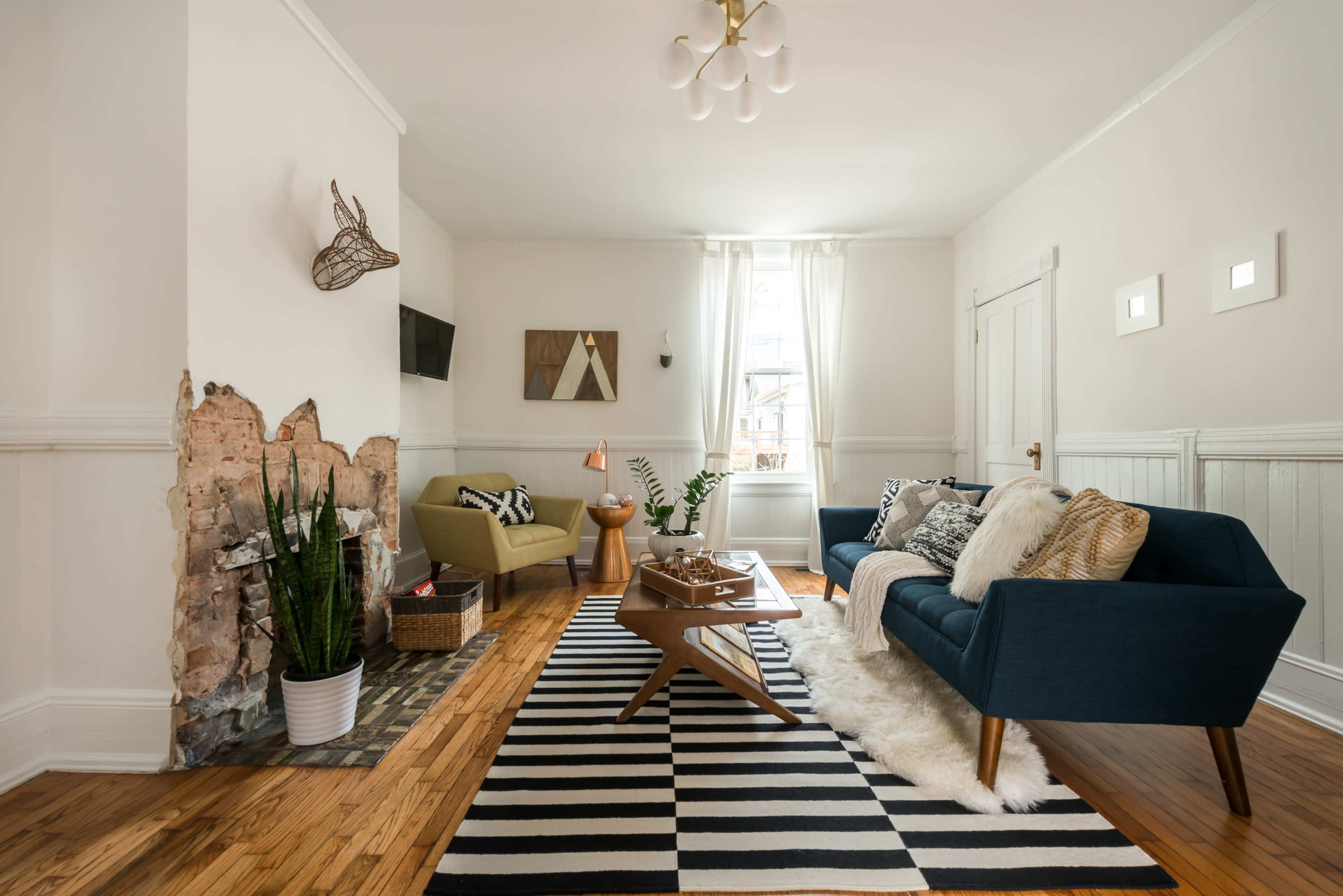 The living room features a blue sofa, a green armchair, a wooden coffee table, and a patterned black and white rug, with a partially exposed brick wall and large windows allowing natural light.