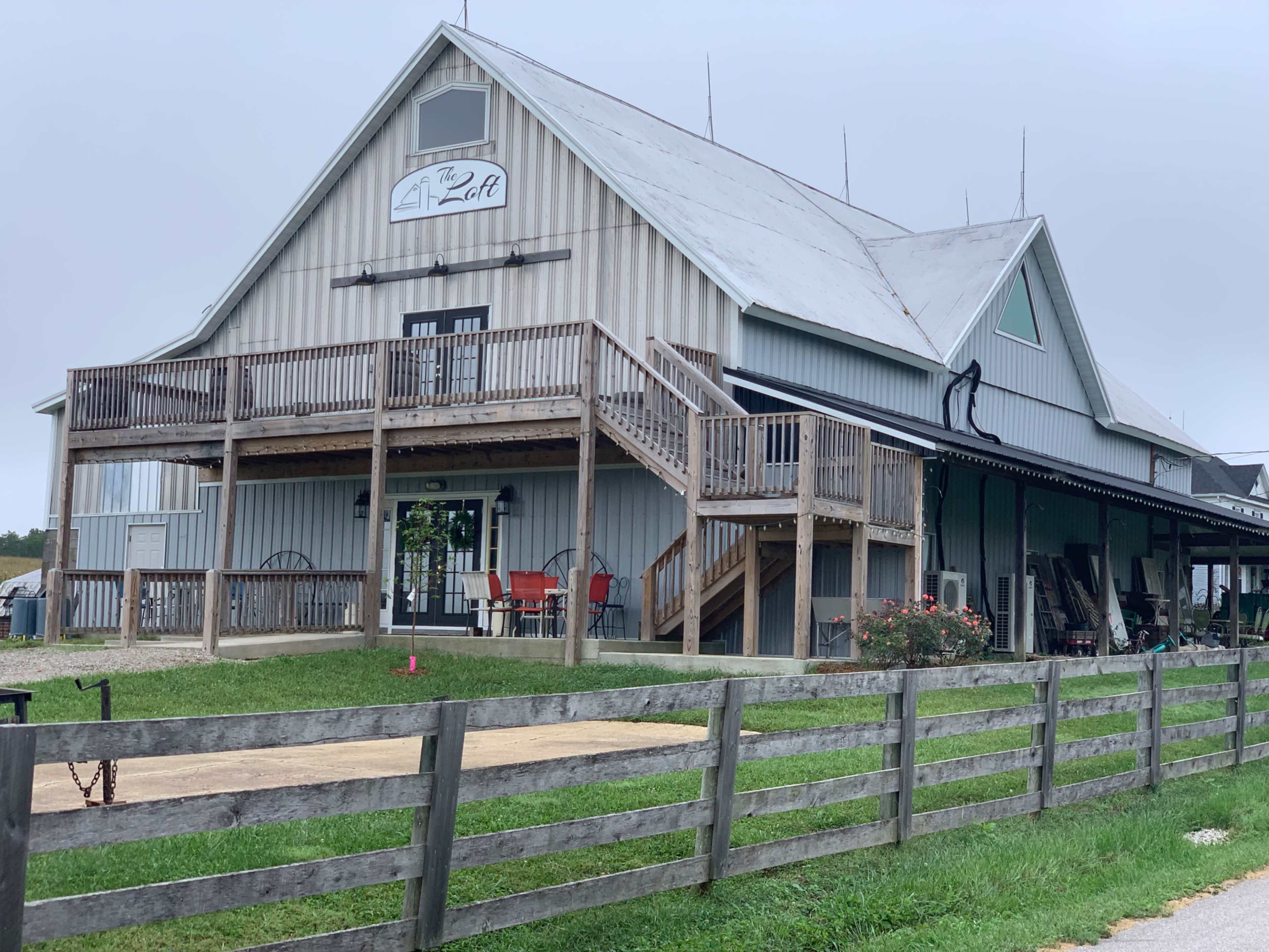 The image shows a large, two-story building with a peaked roof, wide wooden stairs leading to a porch, and a grassy area in front, surrounded by a wooden fence.