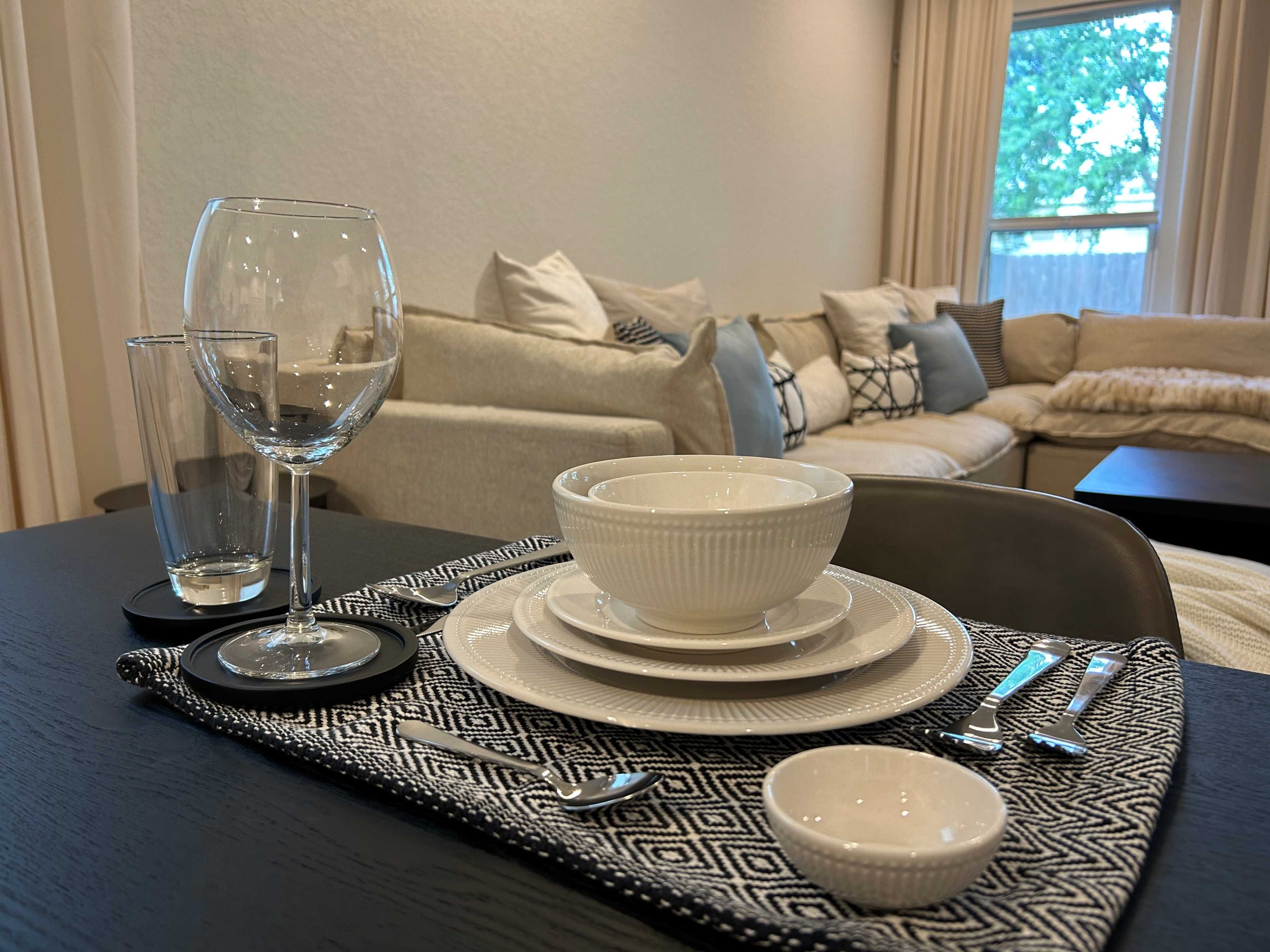 A neatly arranged dining table setting features white dinnerware, glassware, and cutlery, with a cozy living area in the background.