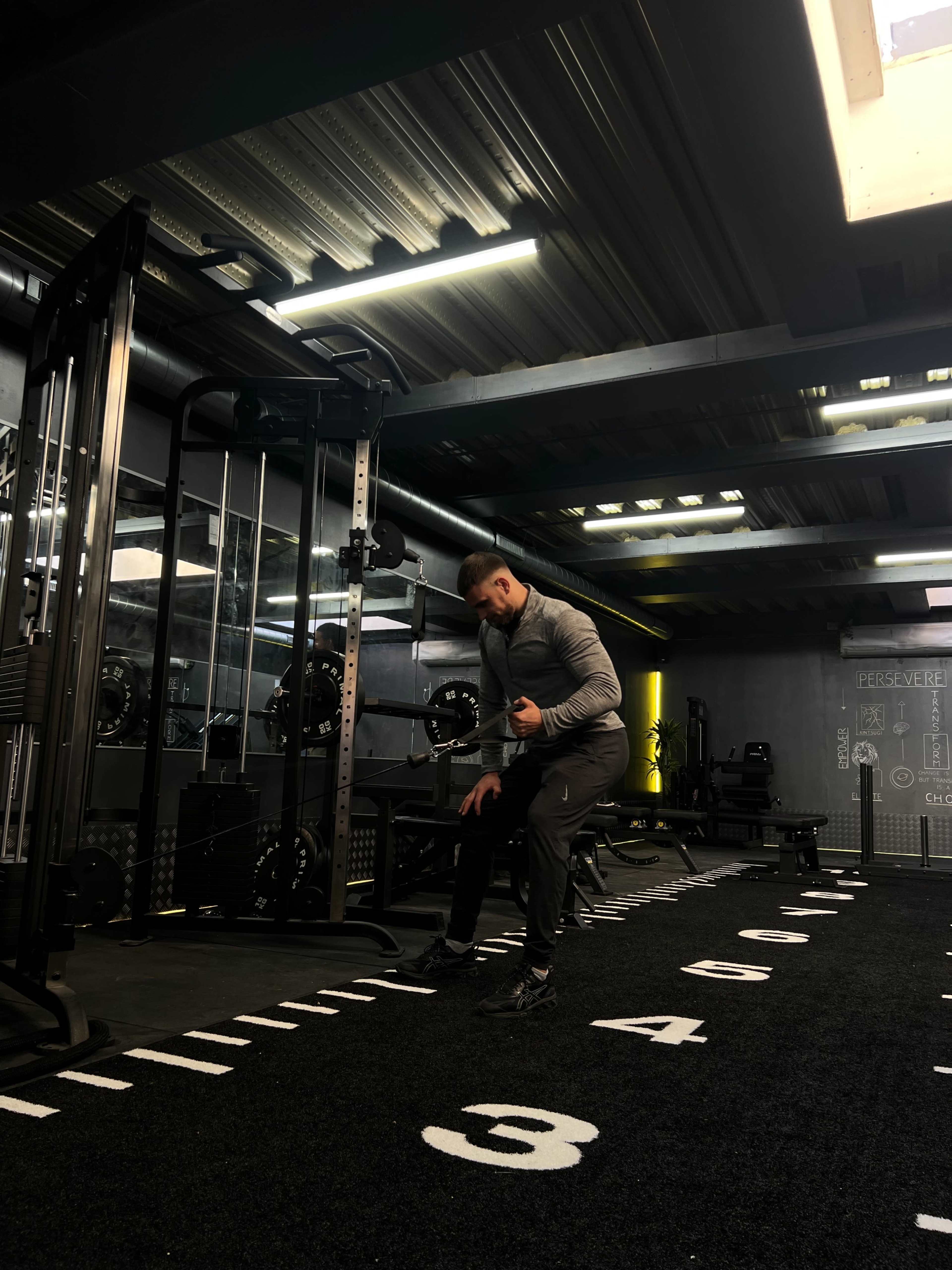 A man is stretching in a gym filled with weightlifting equipment.