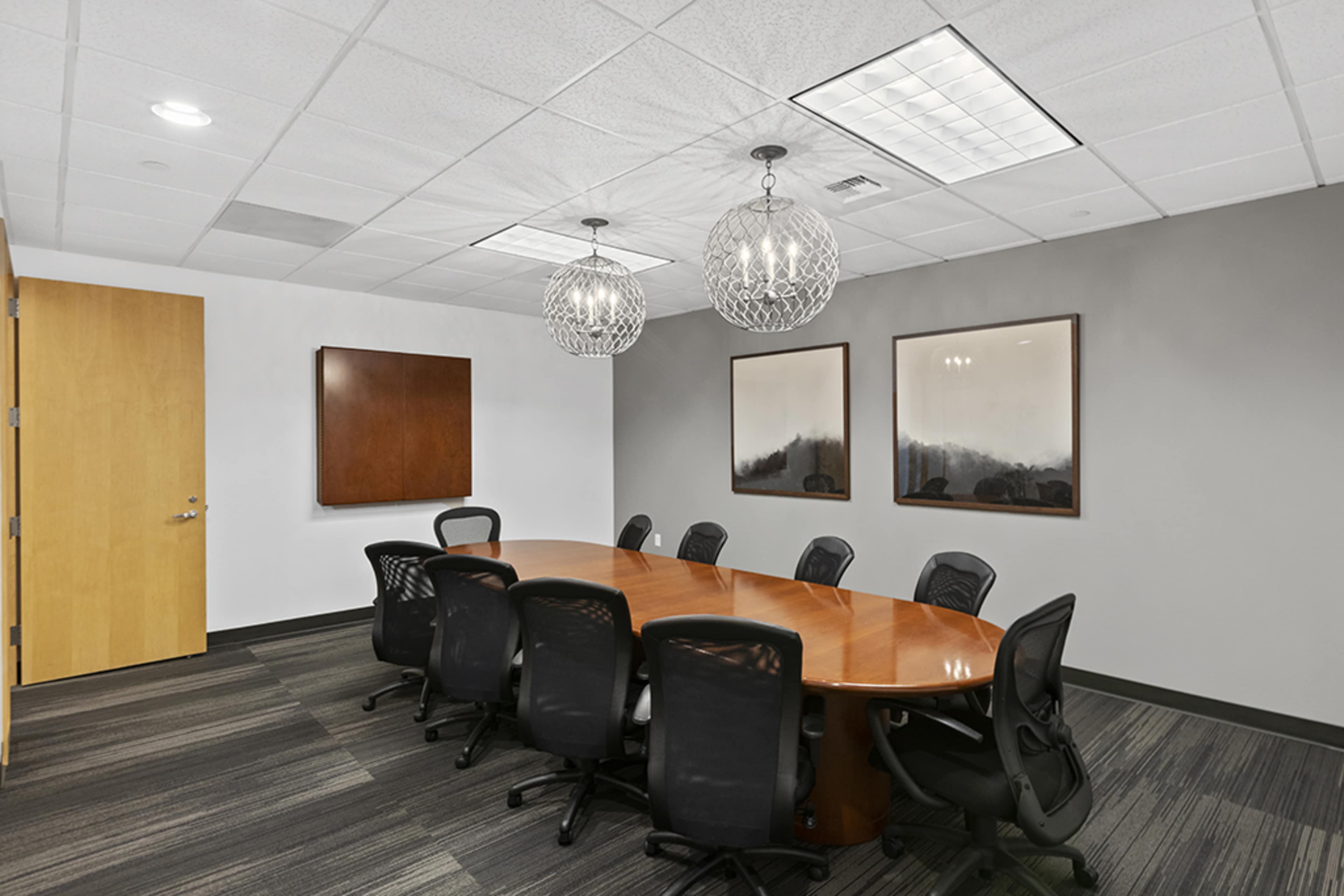 A modern conference room with a large oval wooden table surrounded by black mesh chairs, and two decorative pendant lights hanging from the ceiling.