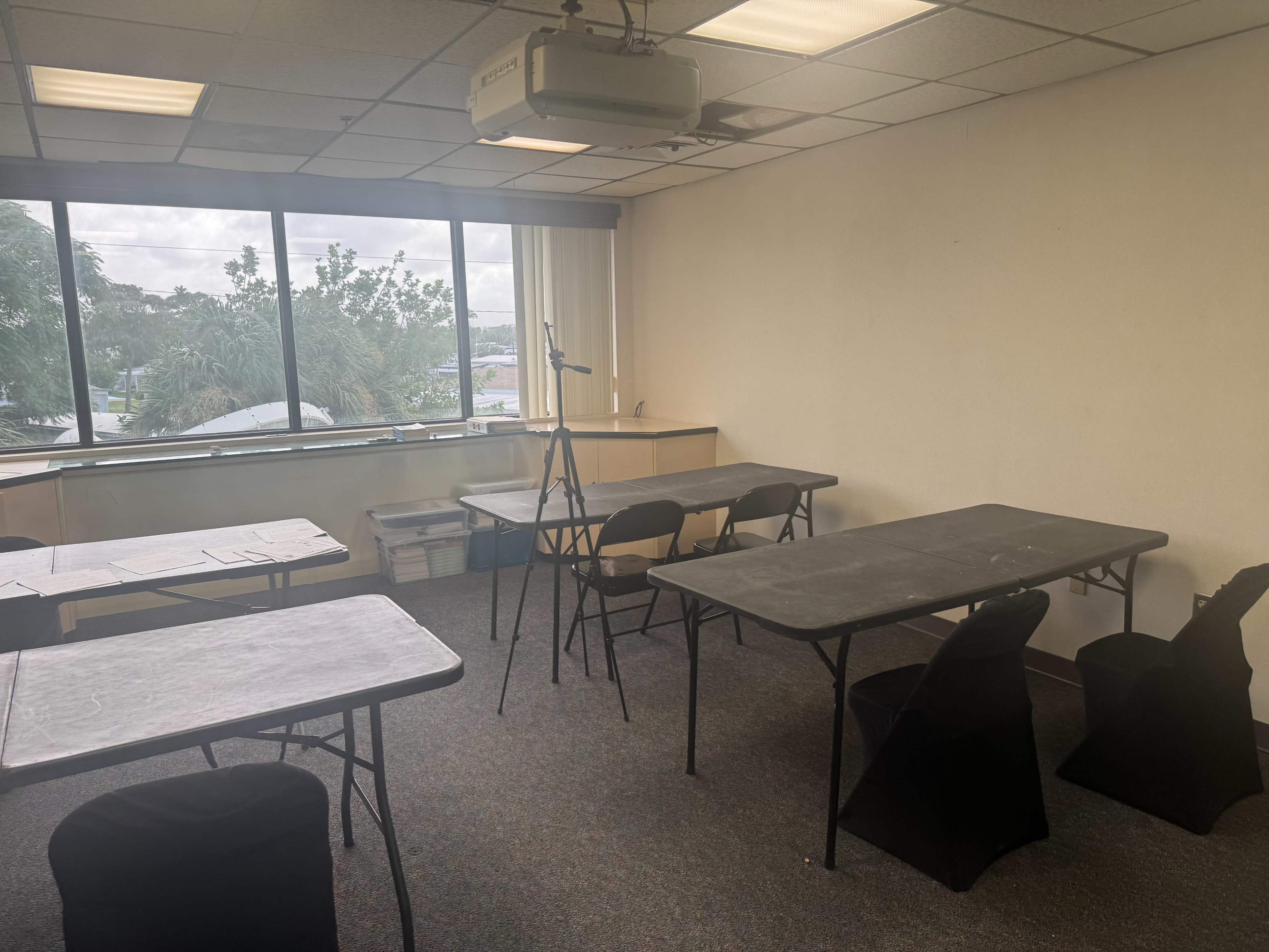 The image shows a sparsely furnished classroom with several tables and chairs arranged, large windows providing natural light, and a projector mounted on the ceiling.