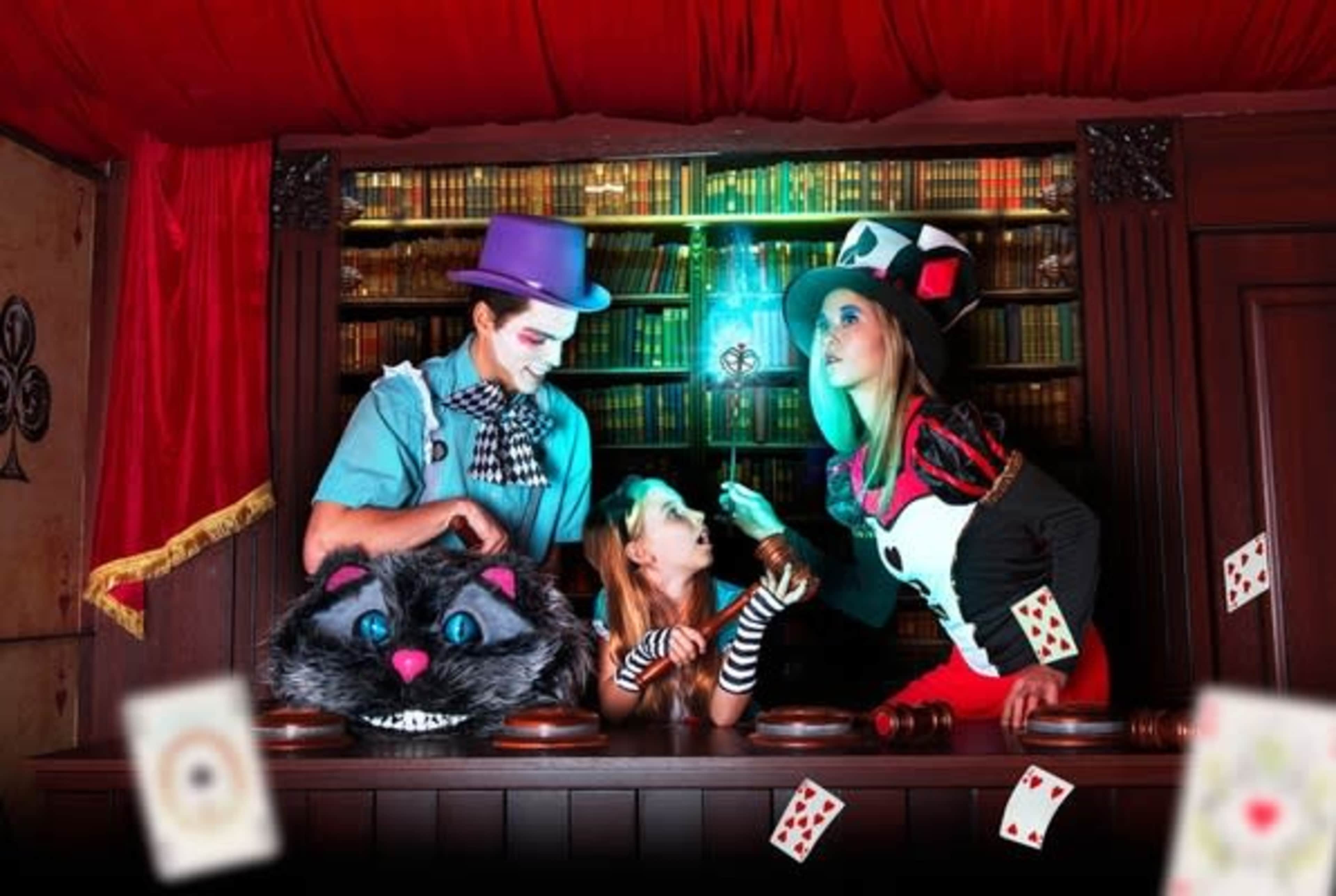 Three individuals dressed in whimsical costumes stand behind a wooden counter adorned with books and playing cards, engaged in a playful scene inspired by a fantastical theme.
