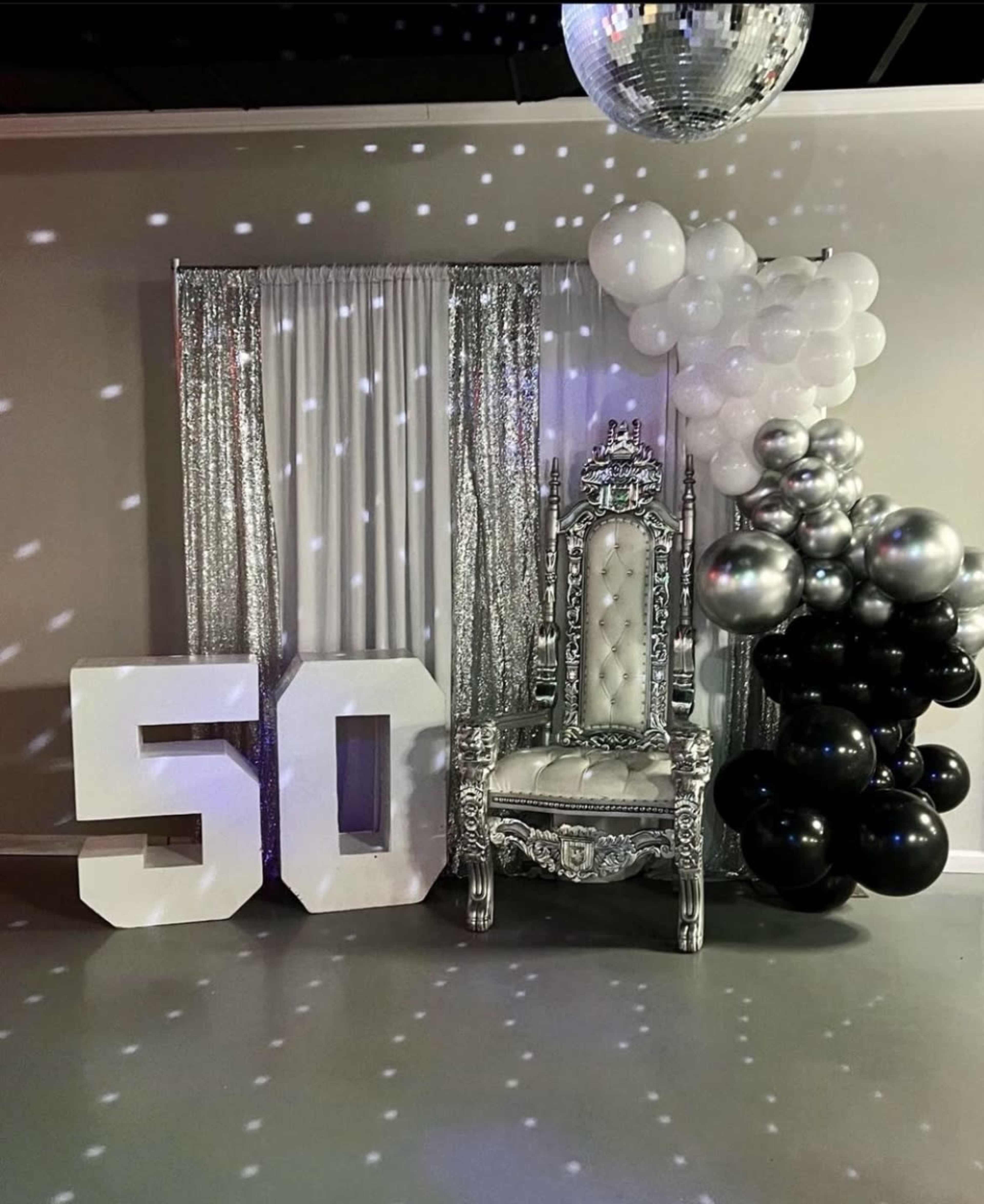 The scene features a decorated area for a 50th birthday celebration, showcasing a large "50" display, an ornate silver throne, and a cluster of black and white balloons.