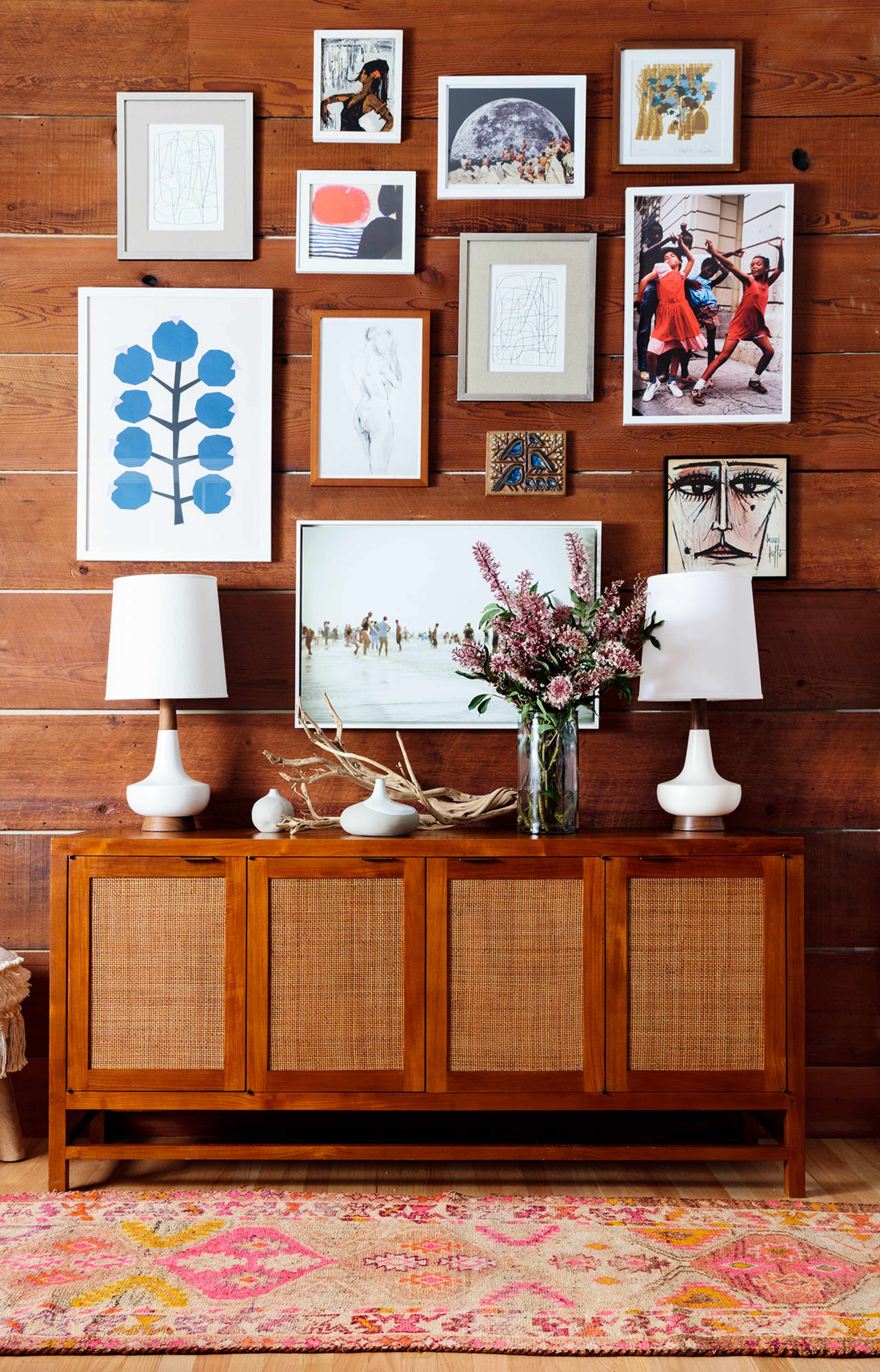 A wooden wall adorned with a collection of framed artworks above a wooden sideboard, which is flanked by two table lamps and displays a vase of flowers.