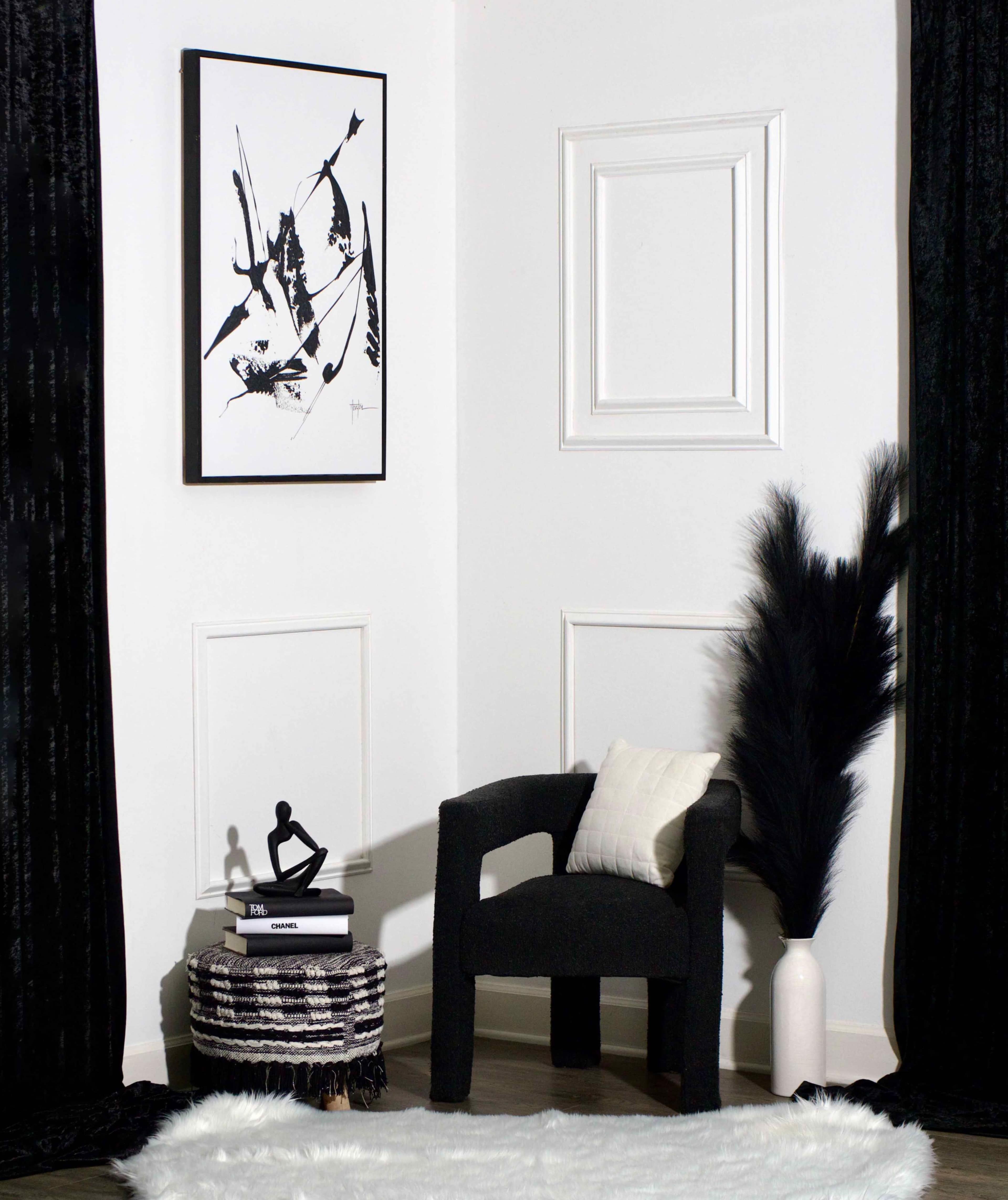 The image shows a corner of a room featuring a black chair, a decorative stool, a white vase with black pampas grass, and an abstract black and white artwork on the wall.