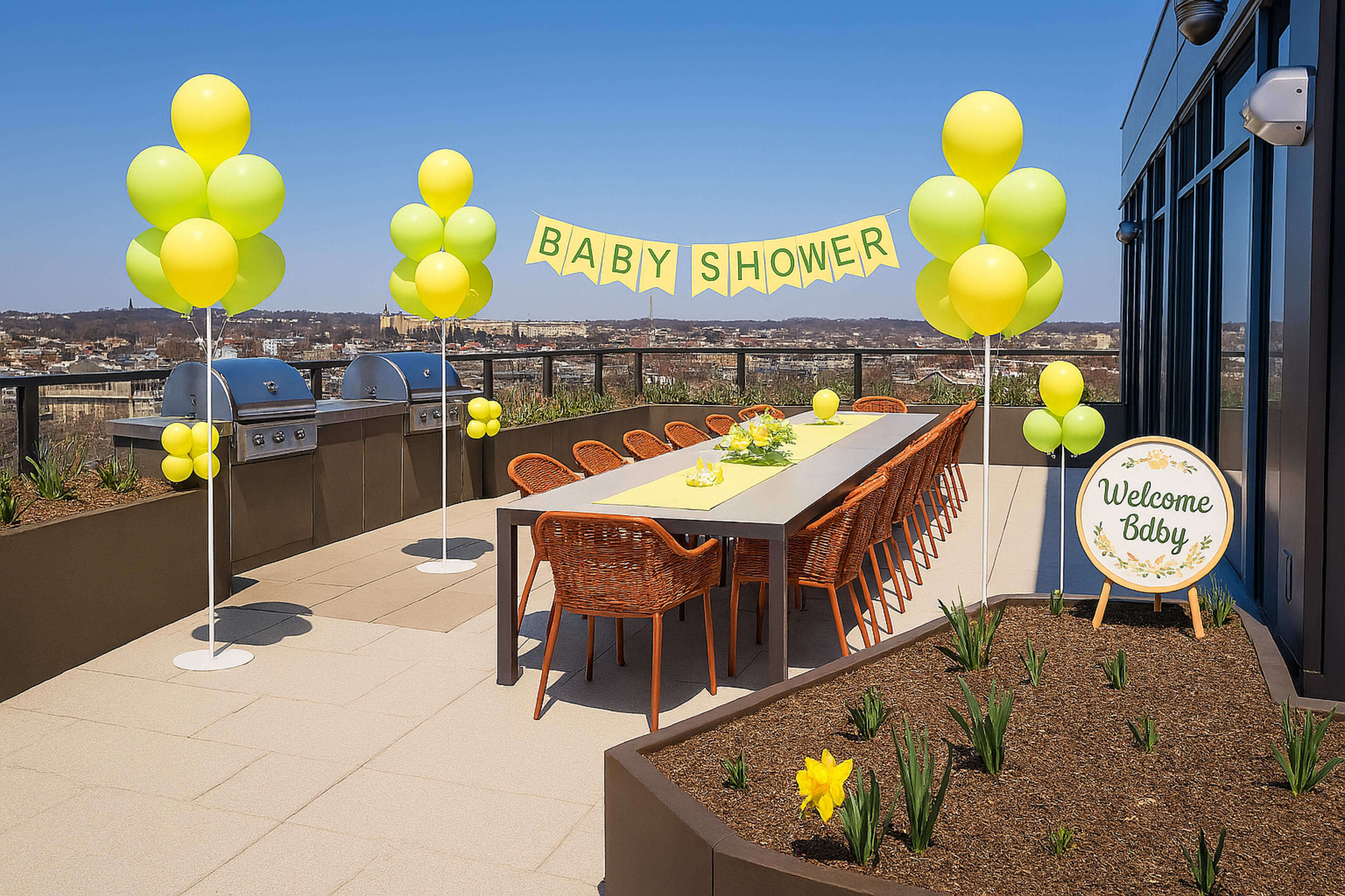 The image shows a rooftop terrace set up for a baby shower, featuring a long table with chairs, yellow and green balloons, and a welcome sign among decorative plants.
