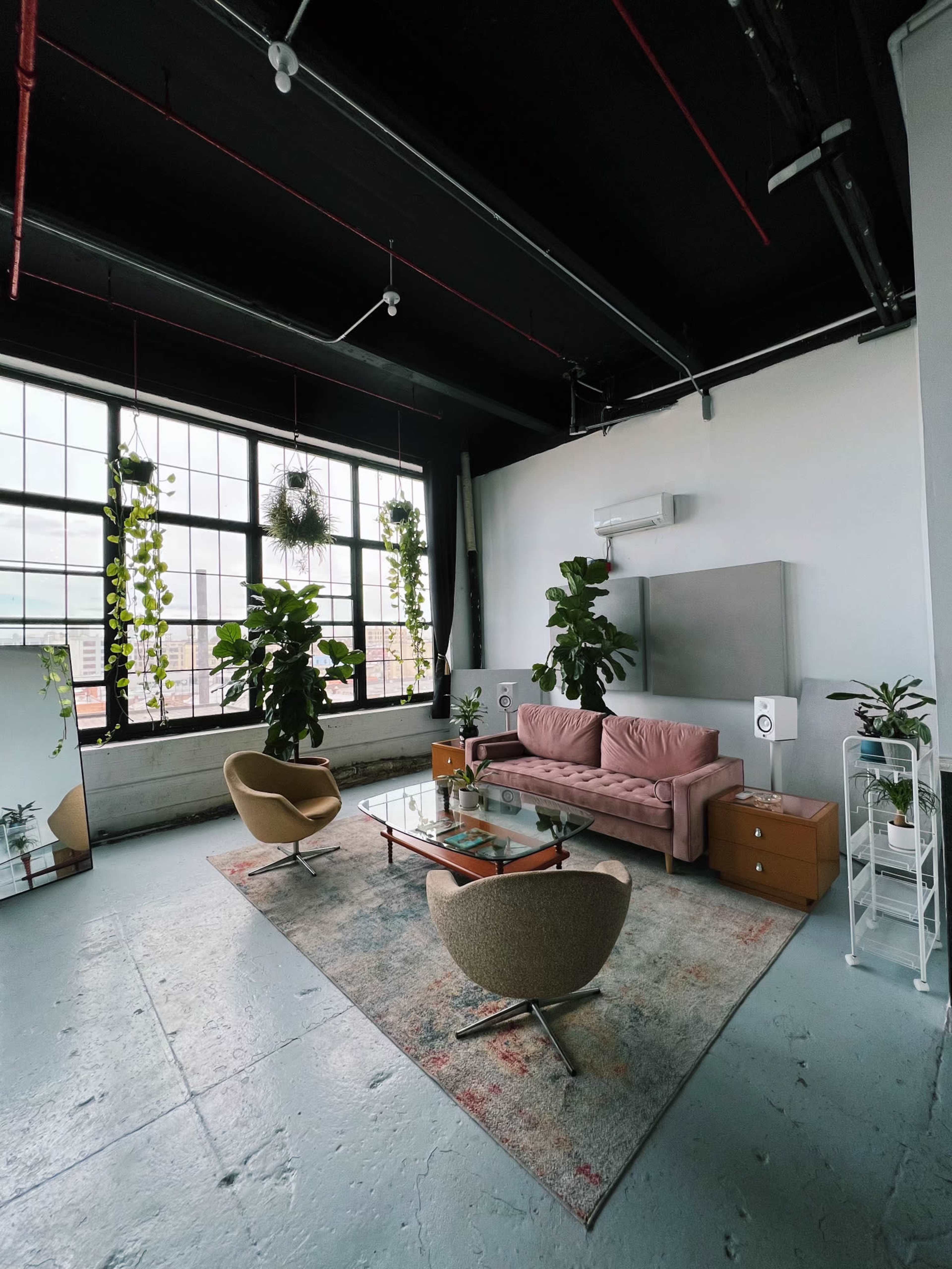 A modern living room featuring a pink sofa, two tan armchairs, indoor plants, a glass coffee table, and large windows with city views.