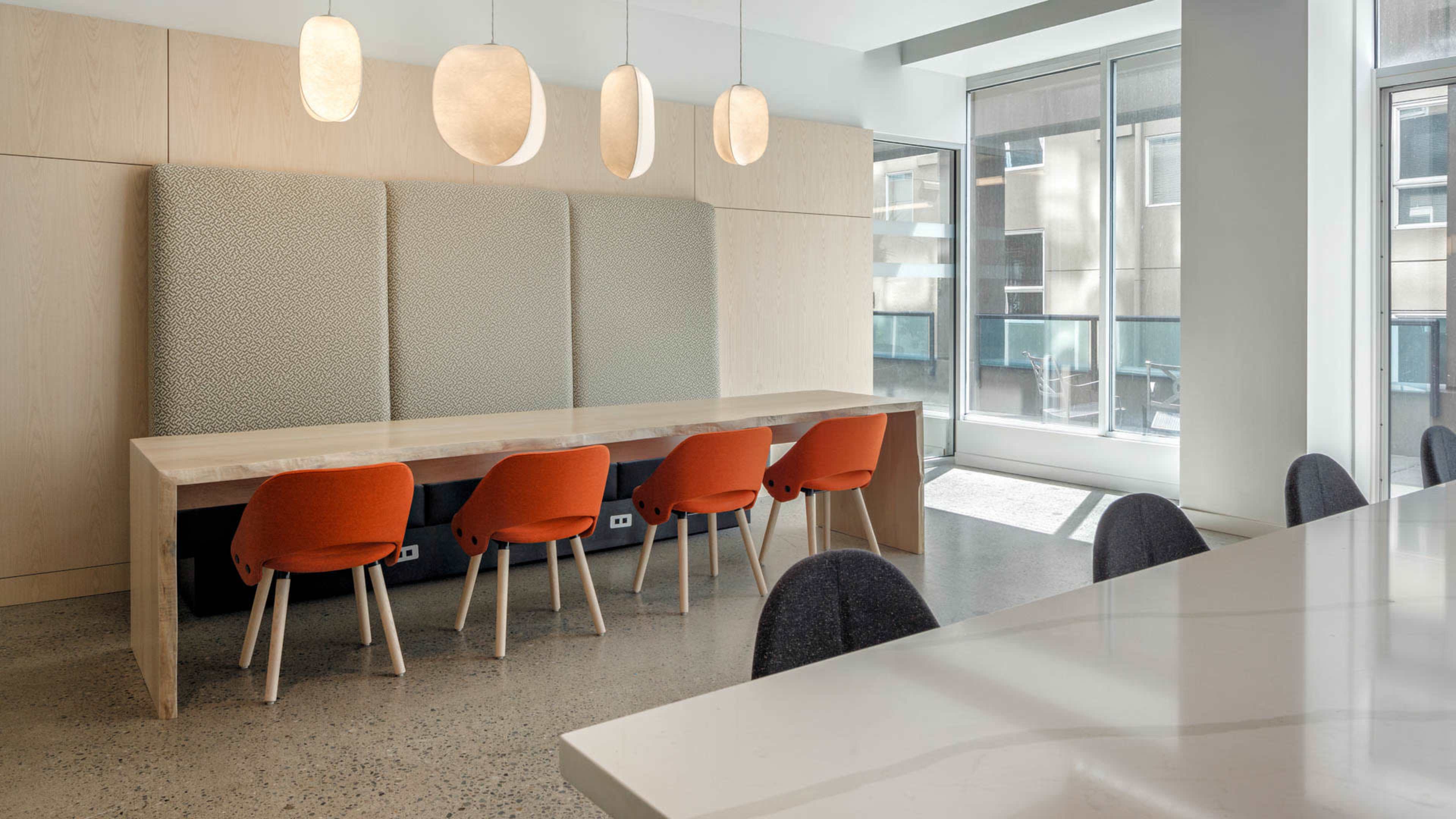A modern dining area features a long wooden table with five orange chairs, complemented by pendant lights and a textured wall backdrop.
