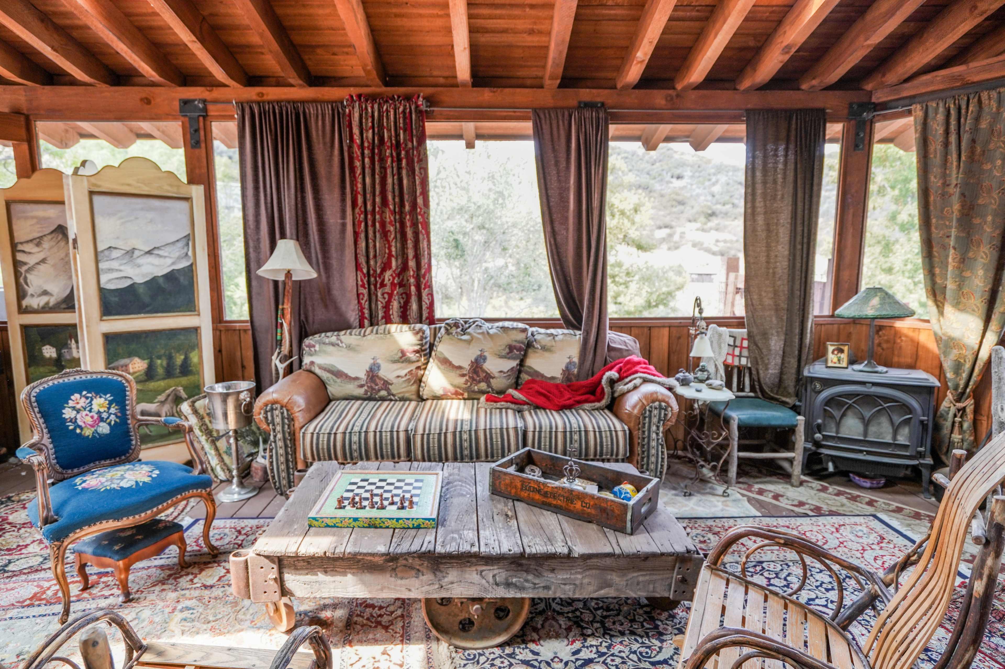 A cozy living room features a leather sofa, a vintage armchair, a wooden coffee table, and an old-fashioned stove, all set against a backdrop of large windows and rustic decor.