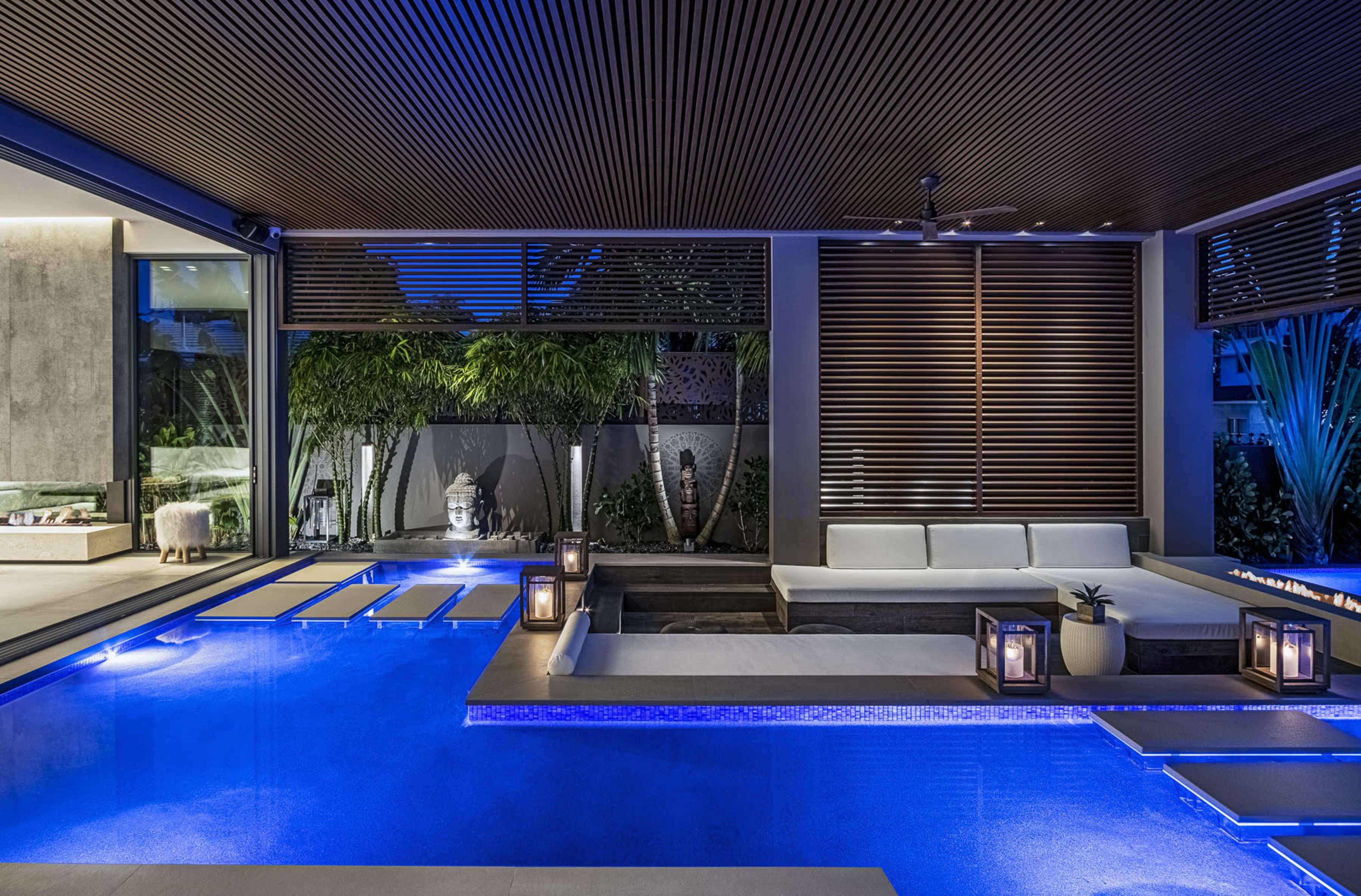 The scene features a modern outdoor lounge area with a swimming pool illuminated by blue lights, surrounded by potted plants and decorative lanterns.