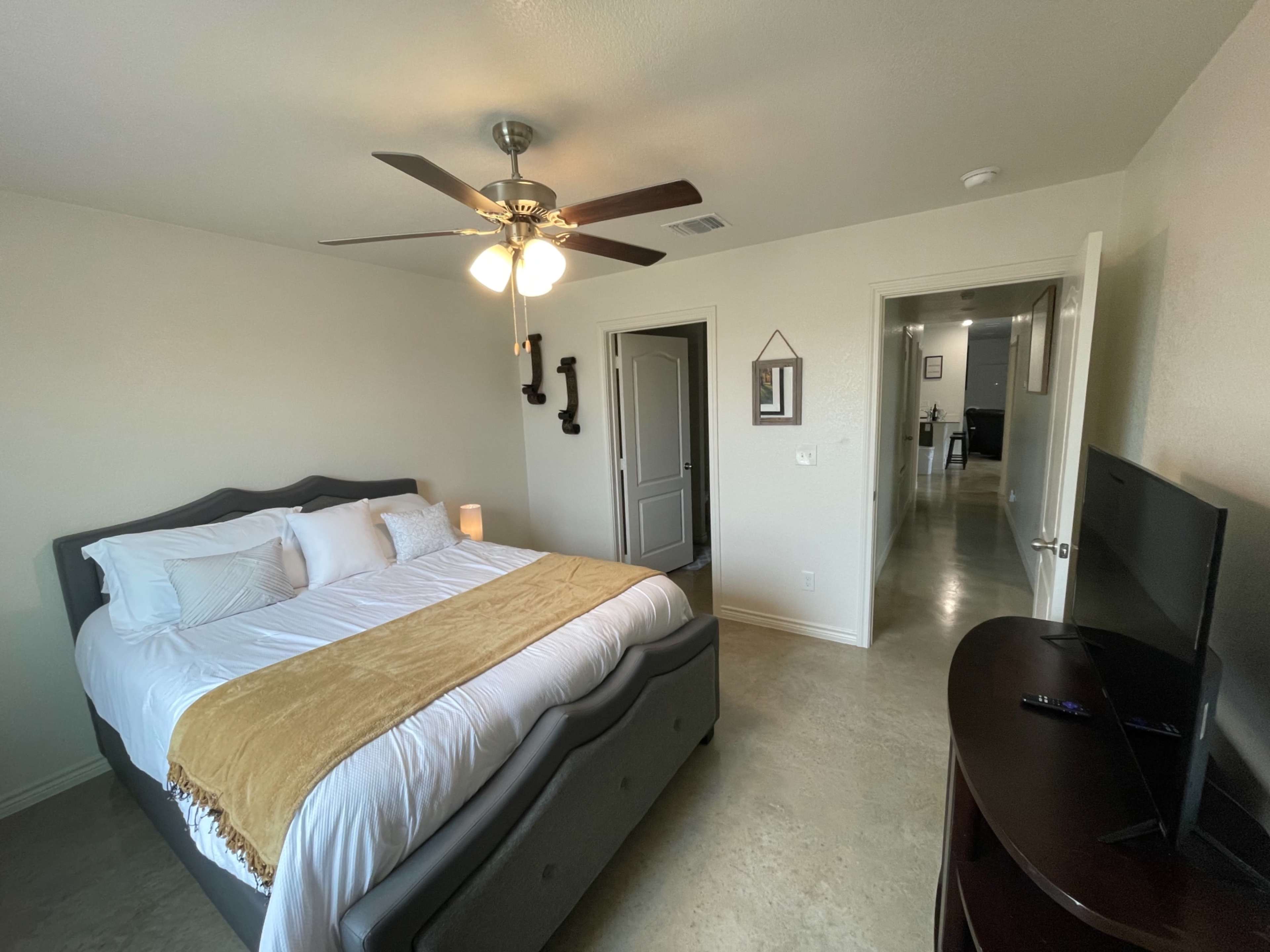 A well-lit bedroom features a king-sized bed with white linens and a gold throw, accompanied by a nightstand, ceiling fan, and a wall-mounted television, with an adjoining hallway visible in the background.