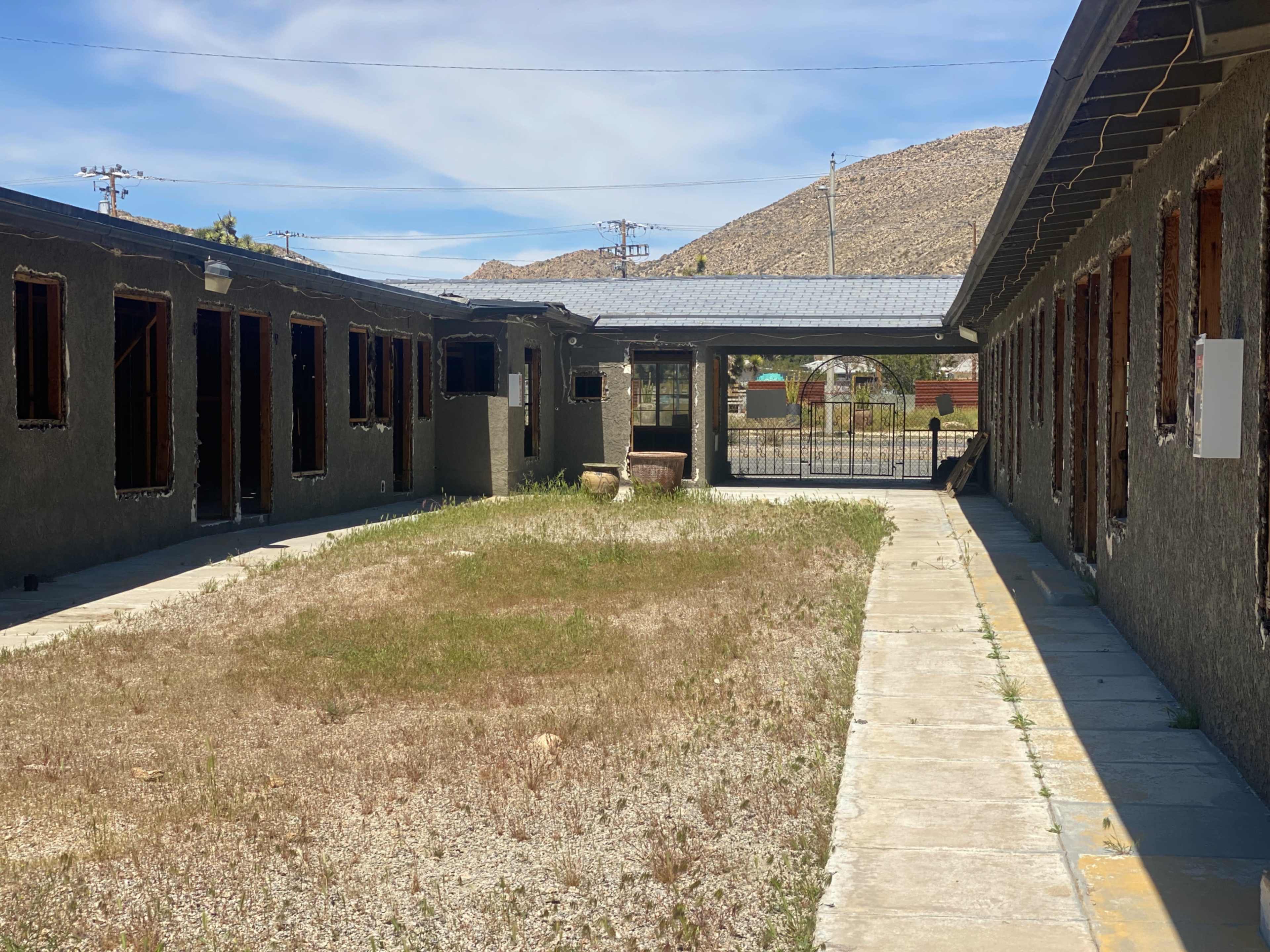 An empty courtyard with cracked pavement and overgrown grass is surrounded by buildings with boarded windows and a mountainous backdrop.