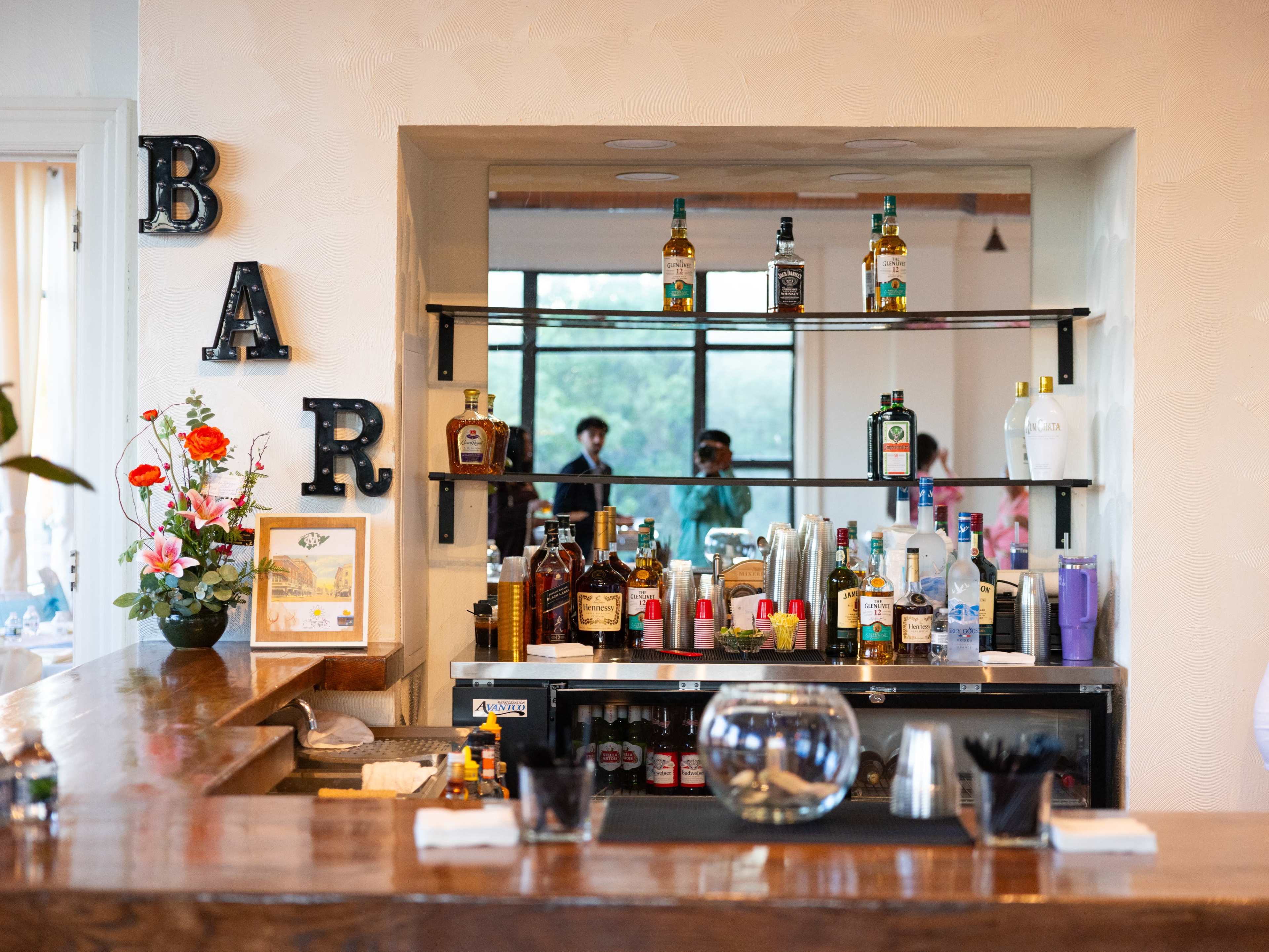 The image shows a neatly organized bar counter with various bottles of liquor displayed on shelves and a reflection of two people in the background.