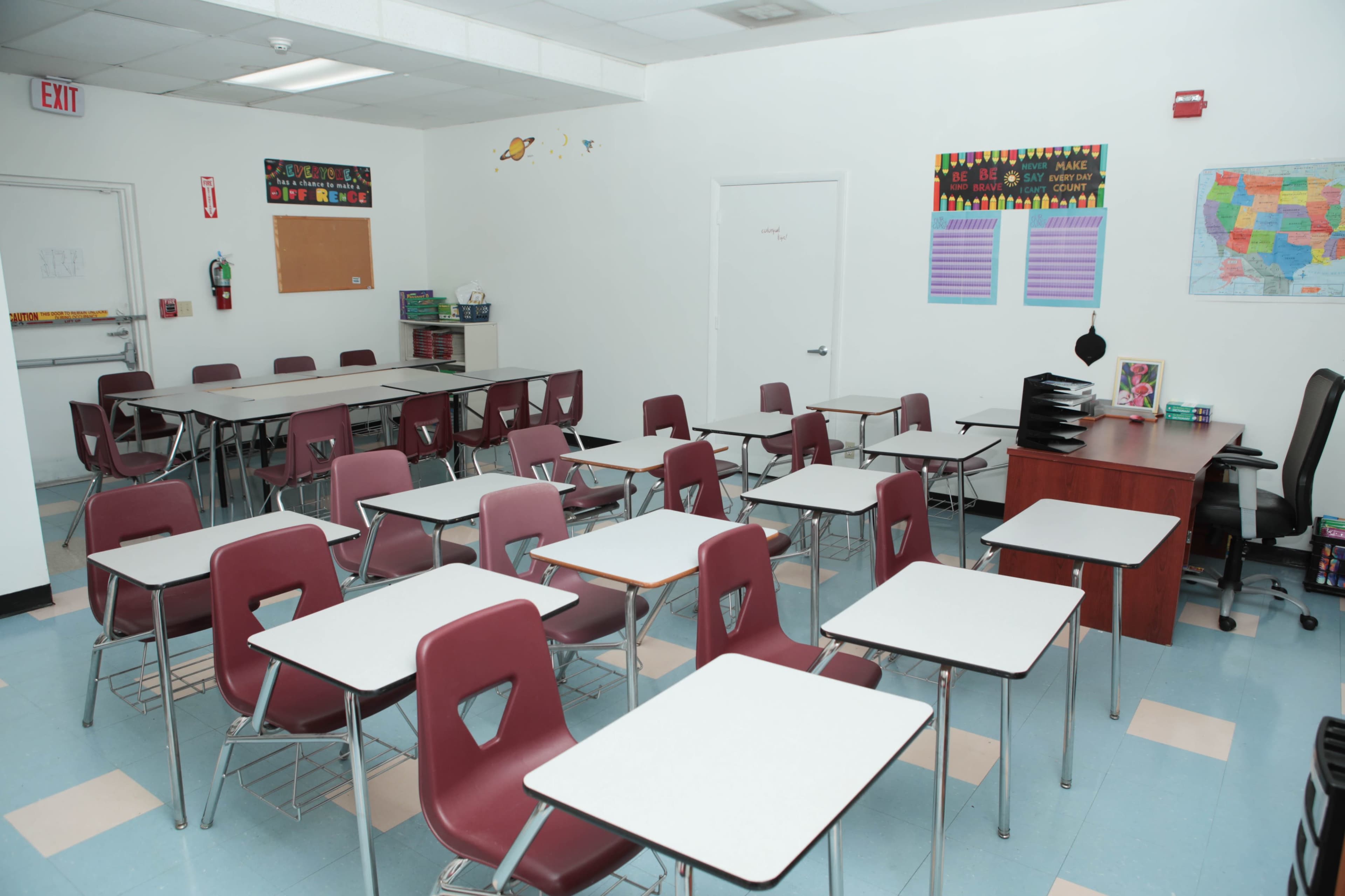 A classroom with rows of metal-framed desks and chairs, a teacher's desk in the corner, and educational posters on the walls.