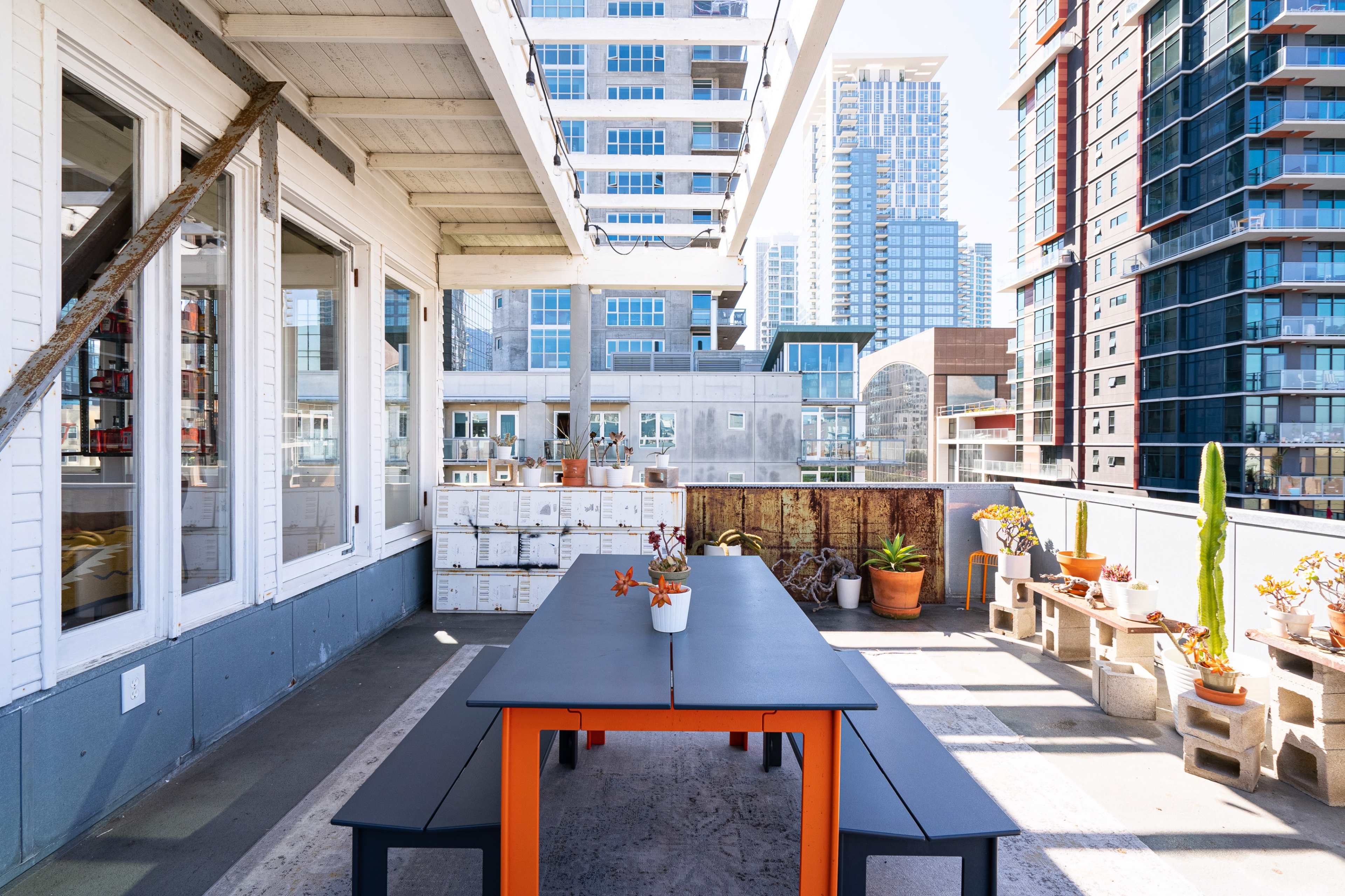 The image shows a rooftop terrace with a black table and benches, surrounded by potted plants and city buildings in the background.
