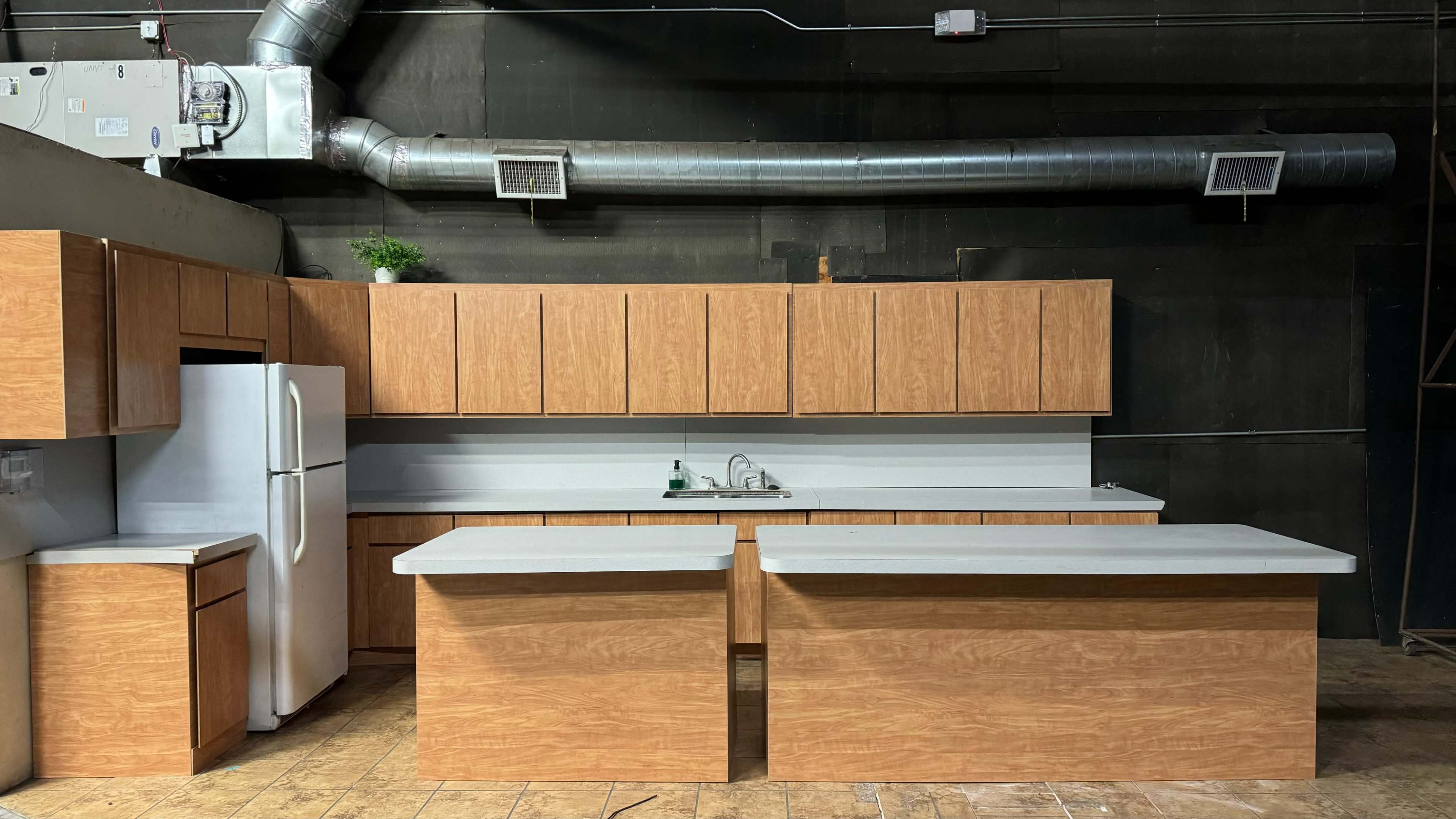 The image shows a modern kitchen setup featuring wooden cabinets, a white countertop, a sink, and a refrigerator against a dark backdrop.