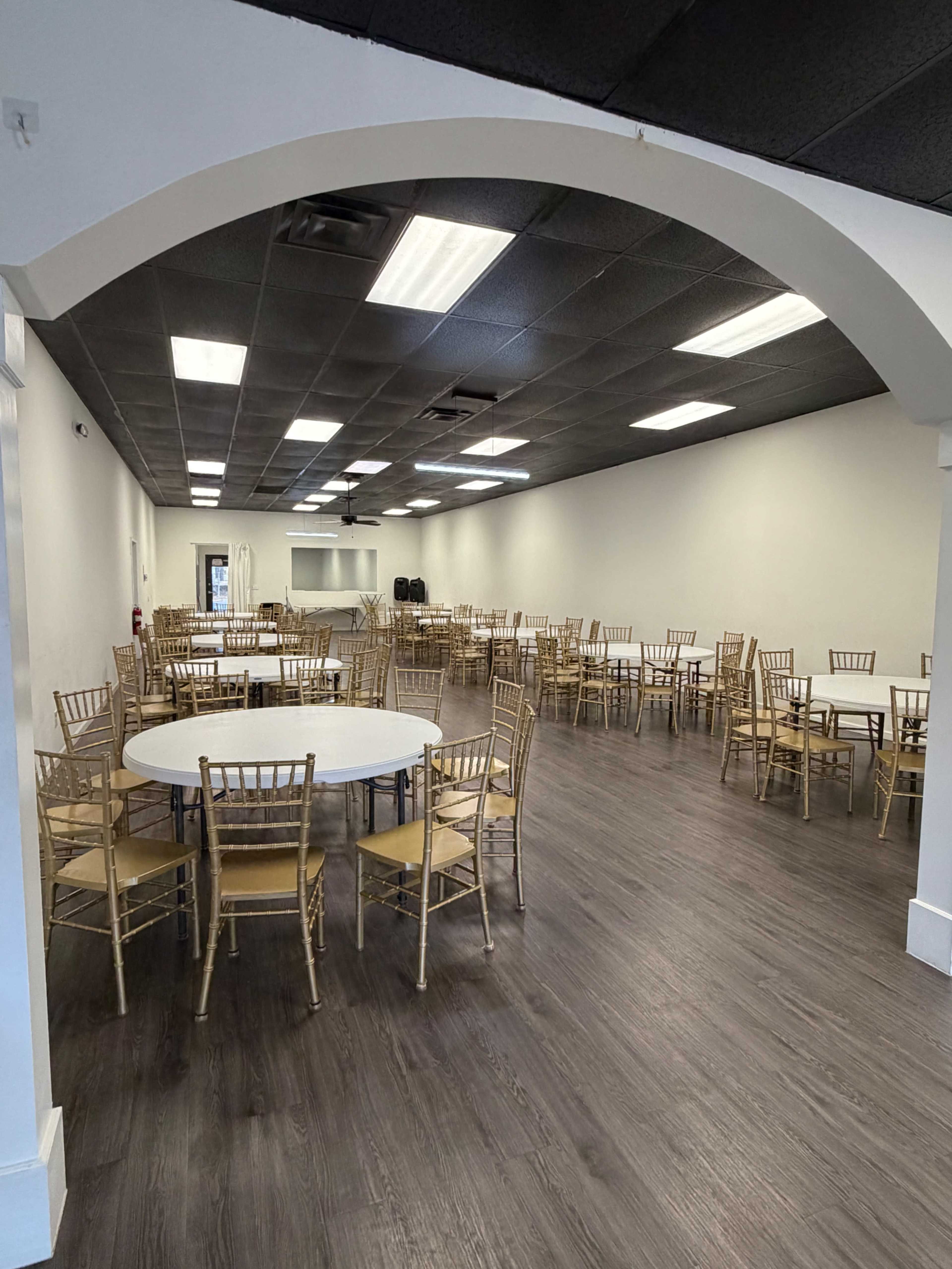 The image shows a spacious banquet hall filled with round tables and gold chiavari chairs arranged in neat lines.