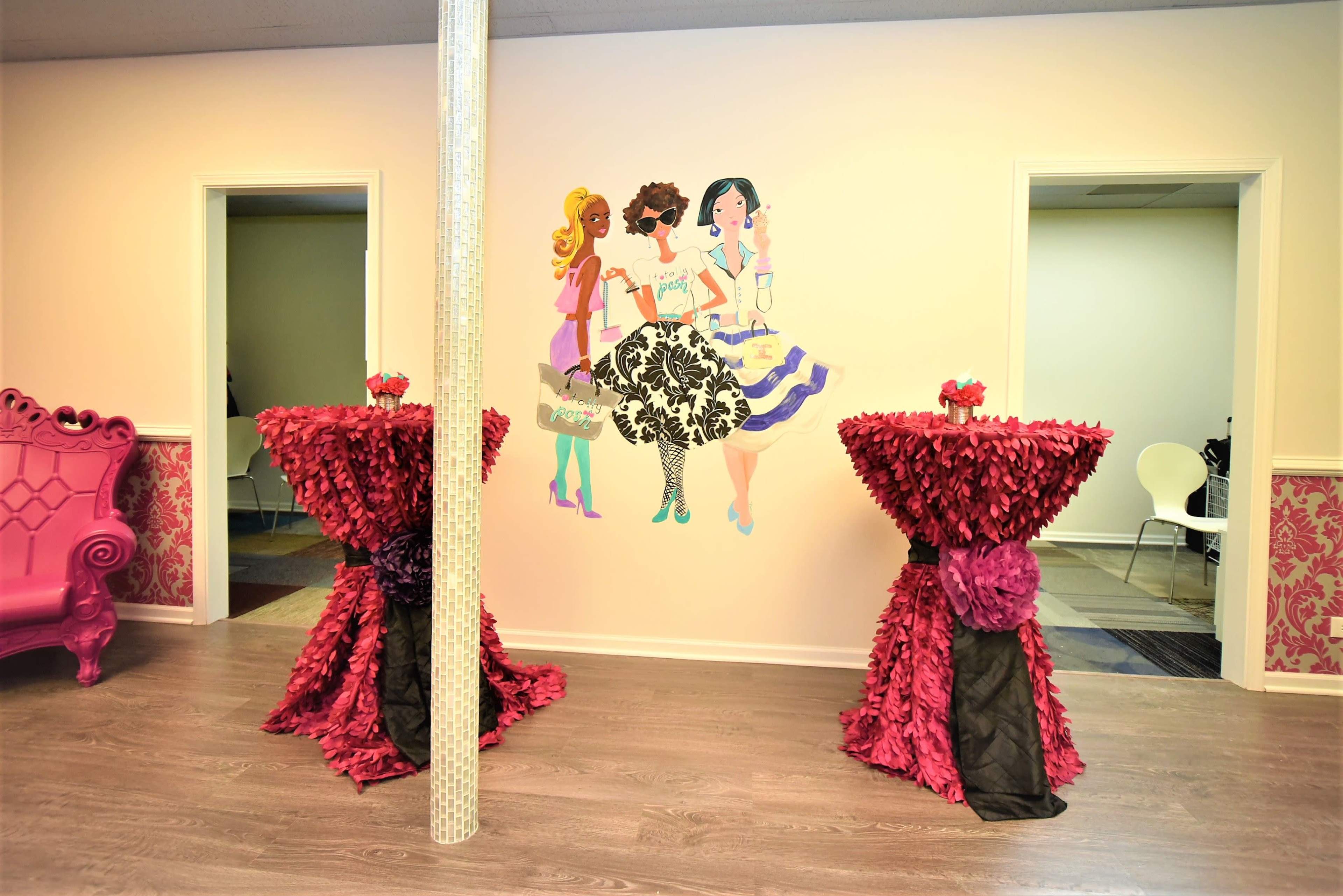 The image features a brightly decorated room with two table displays covered in red ruffles and a wall mural of three fashionable women.