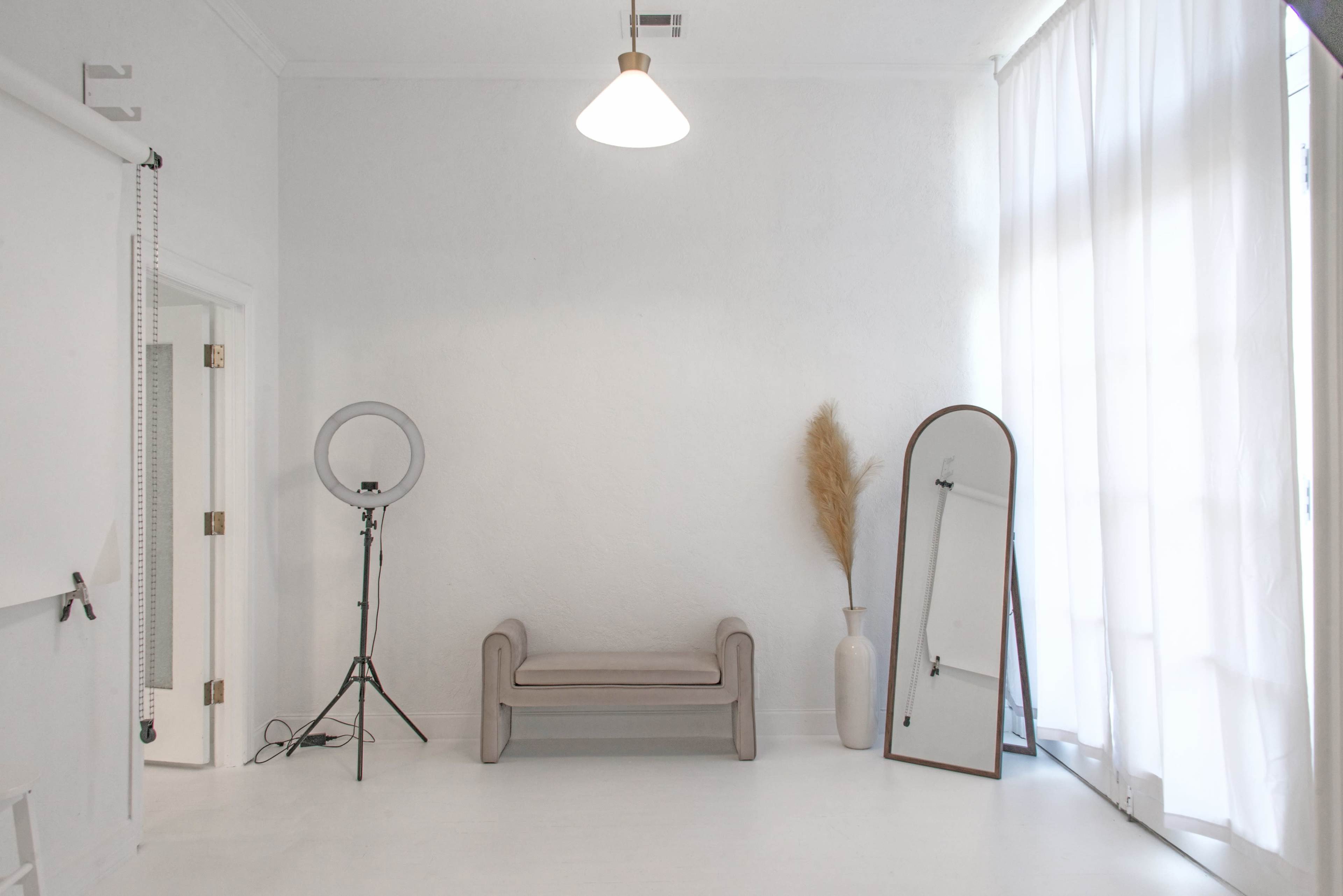 The image shows a minimalist, bright studio interior featuring a light-colored couch, a tall mirror, a floor lamp, and a decorative plant in a vase.