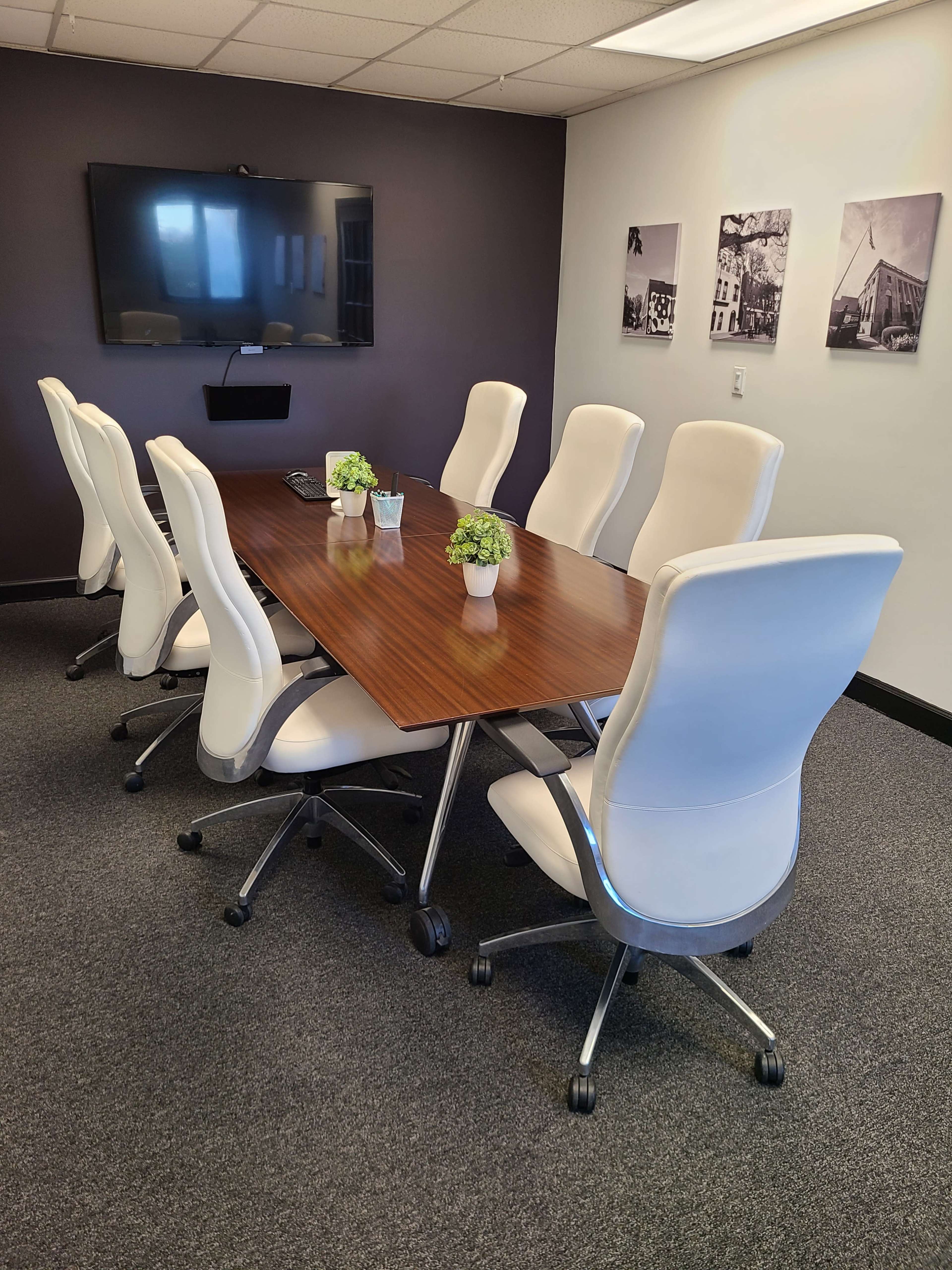 A modern conference room features a large wooden table surrounded by eight white ergonomic chairs and a wall-mounted television.