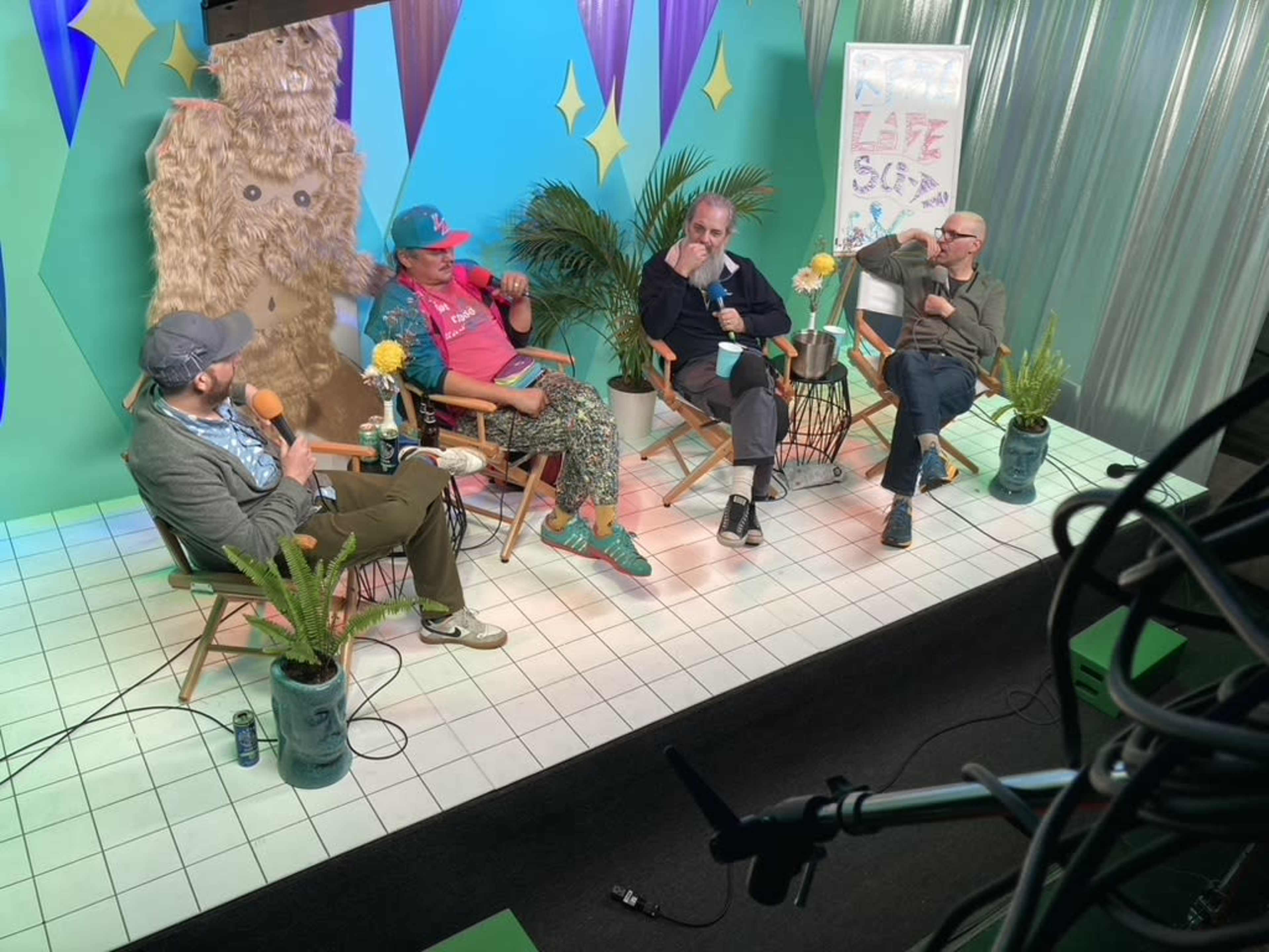 Four individuals are seated in a brightly colored studio setting, engaged in a discussion, with potted plants and a large textured backdrop behind them.