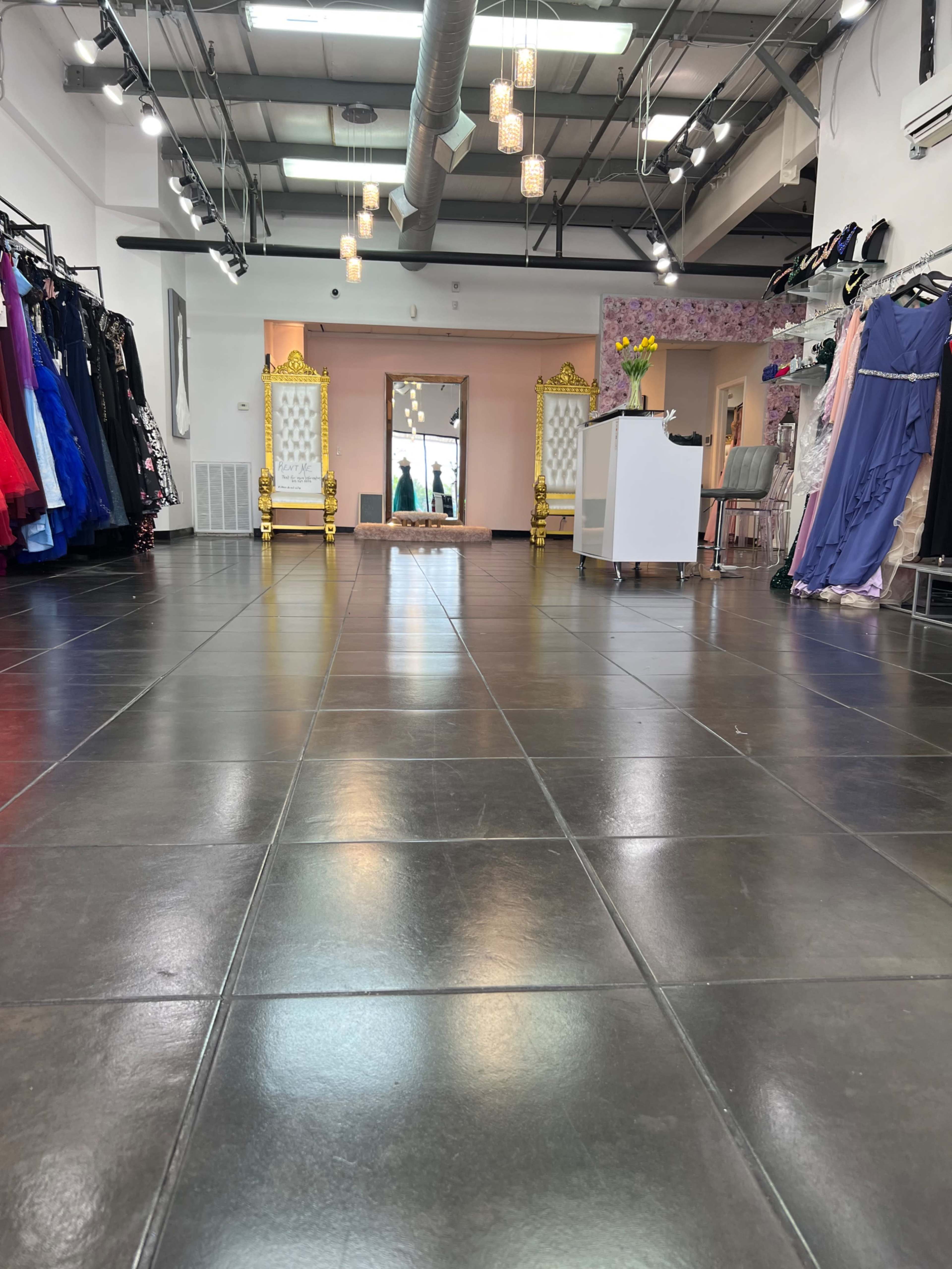A spacious boutique with formal dresses hanging on racks, a large mirror, and decorative golden chairs.