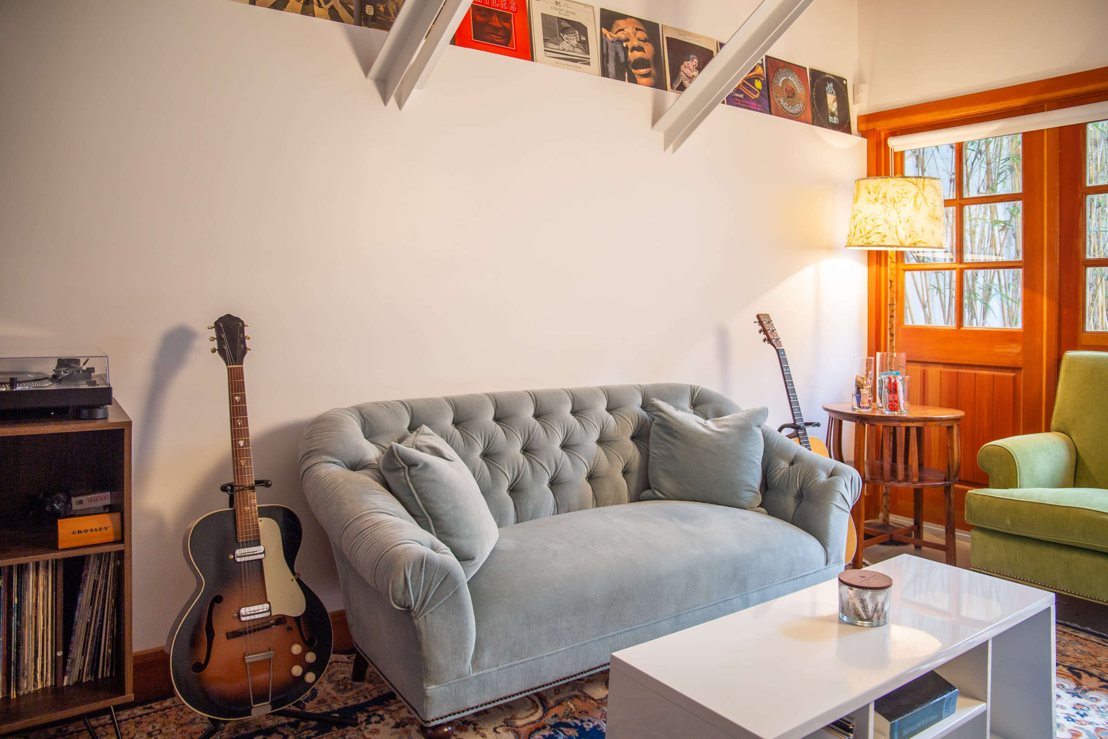 A light blue tufted sofa sits in a living room with a record collection above, accompanied by a guitar and a green armchair.