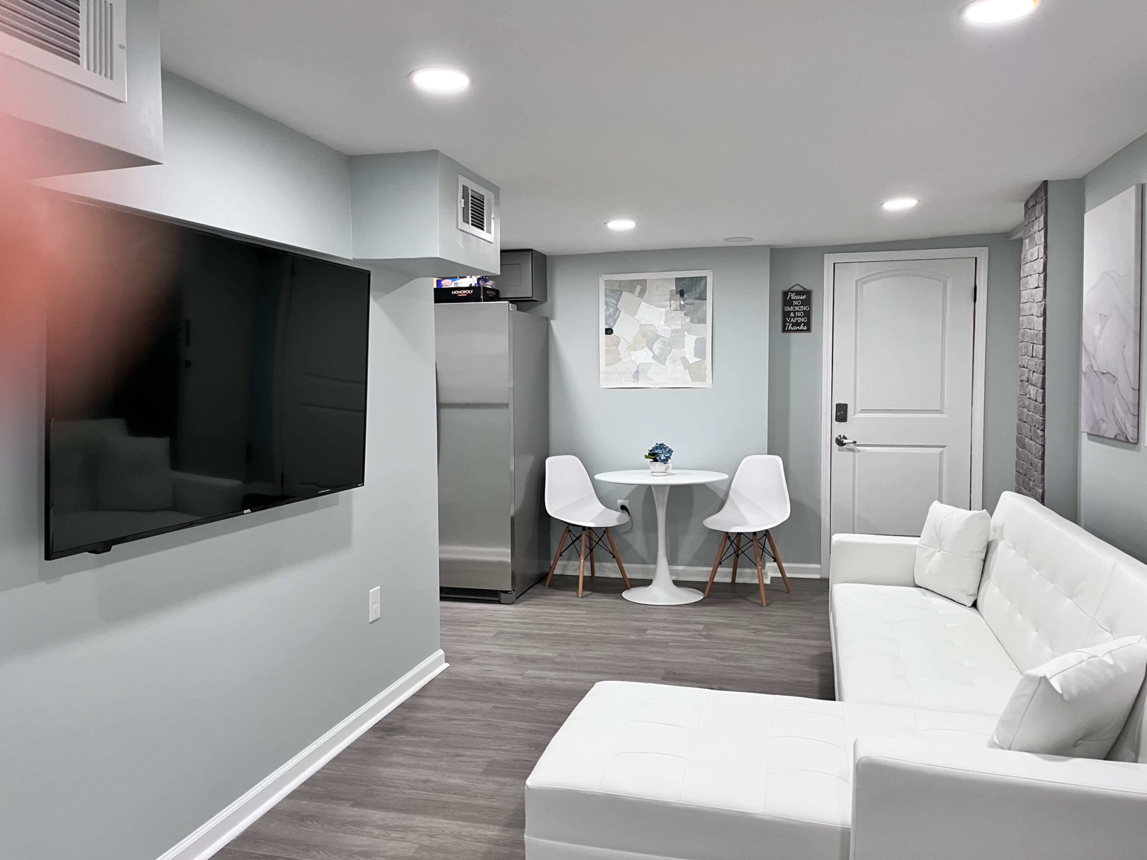 A modern living space features a white sofa, a round dining table with two chairs, a flat-screen television mounted on the wall, and a kitchenette area.