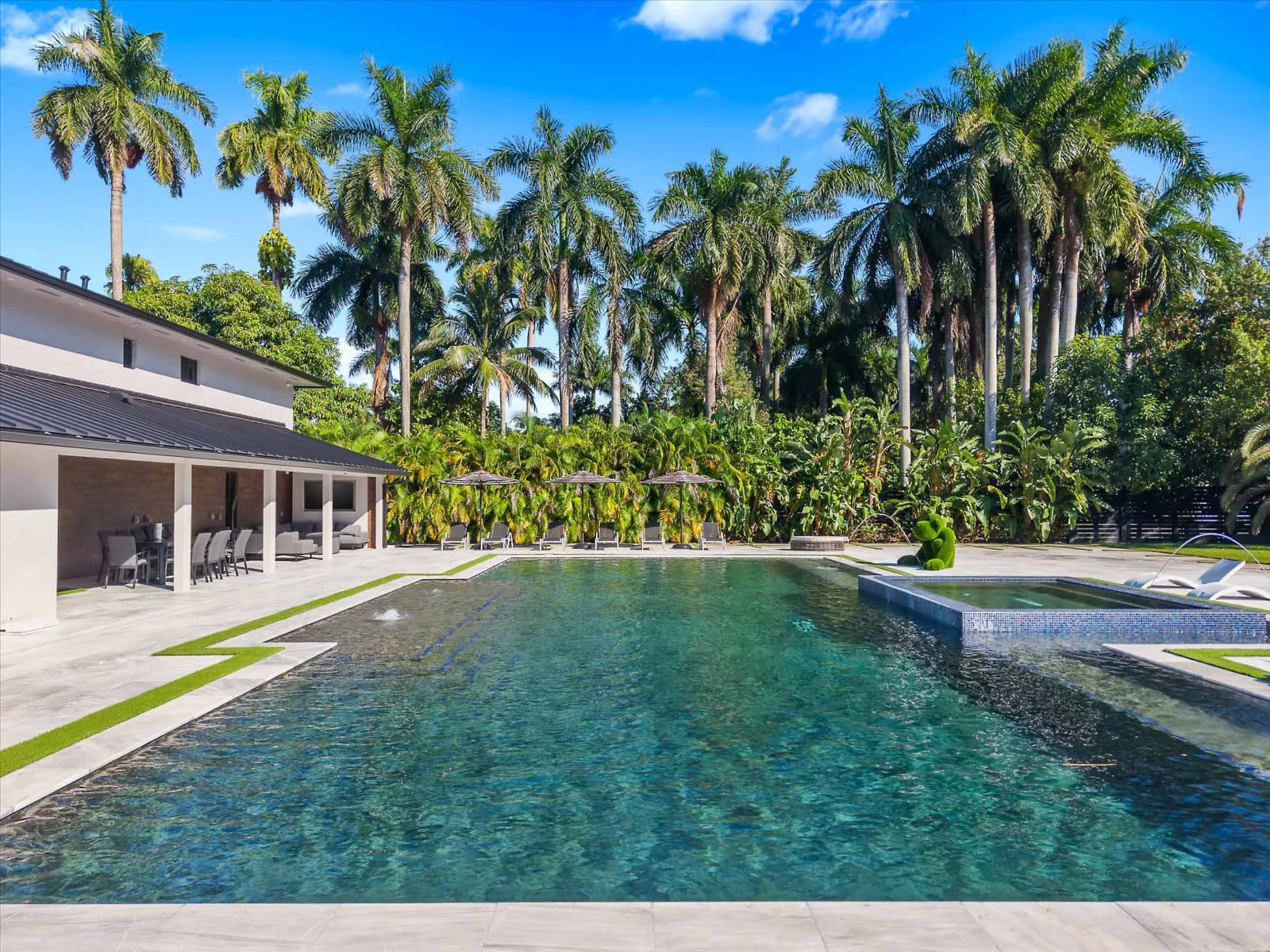 The image shows a large swimming pool surrounded by palm trees and a spacious deck with lounge chairs and umbrellas.