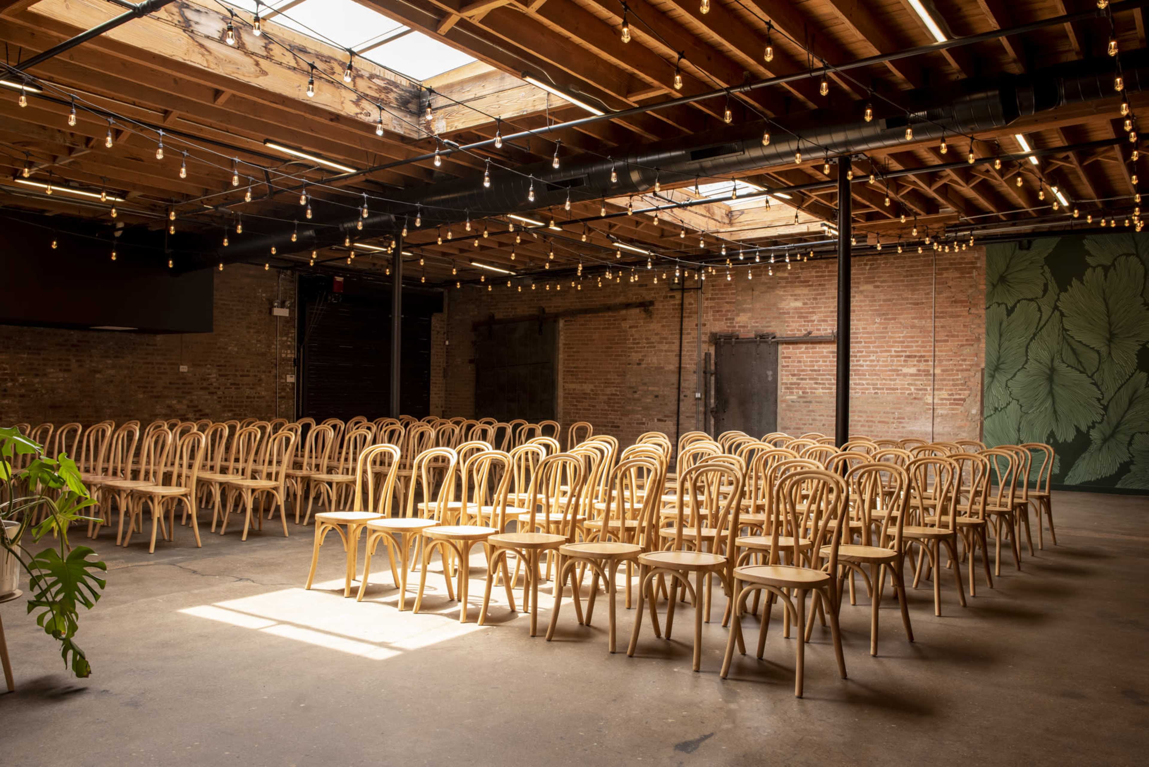 The image depicts a spacious room filled with rows of wooden chairs under a ceiling adorned with string lights.