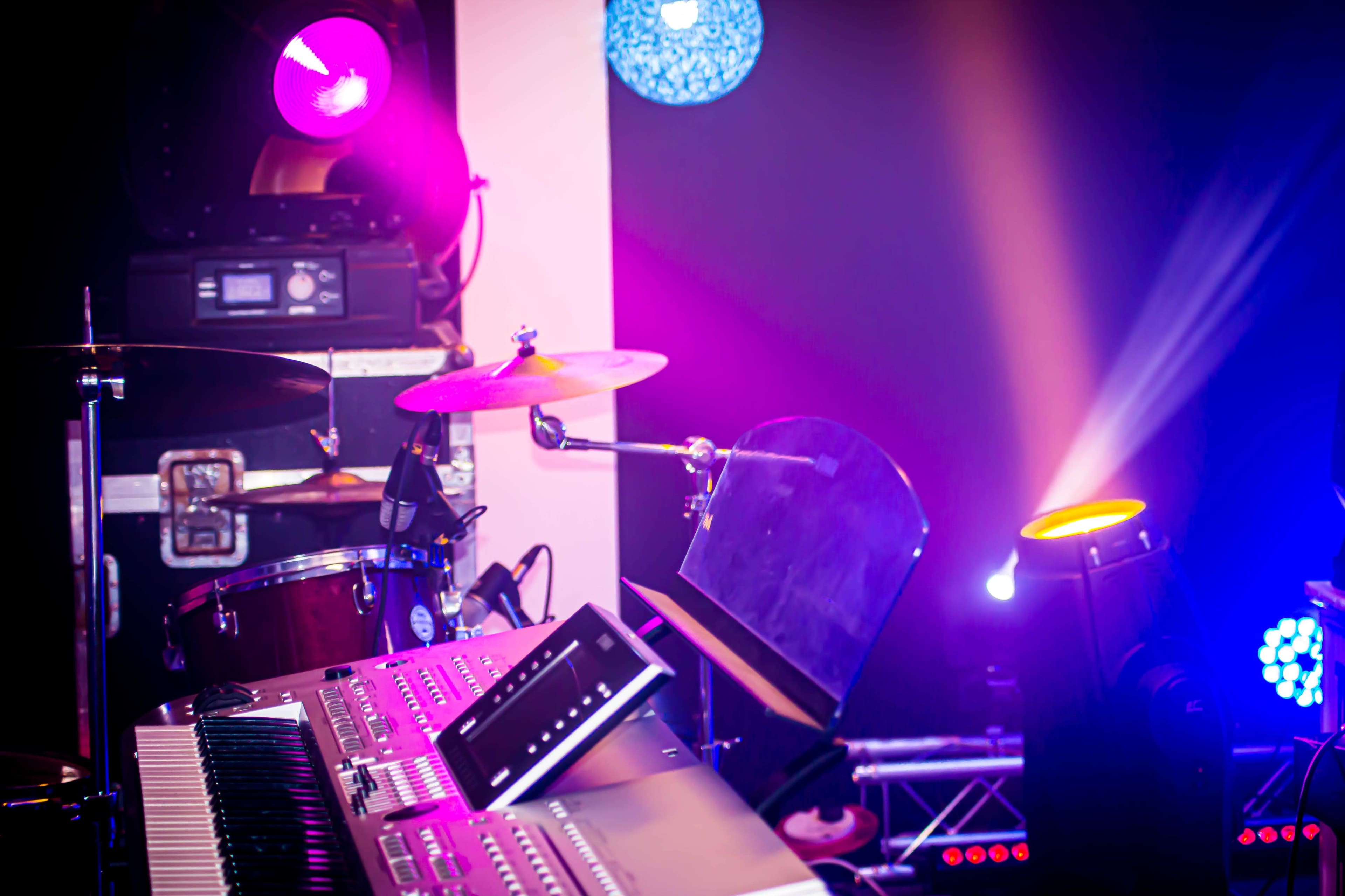 A close-up view of a drum set and a keyboard placed on a stage with colorful lights in the background.