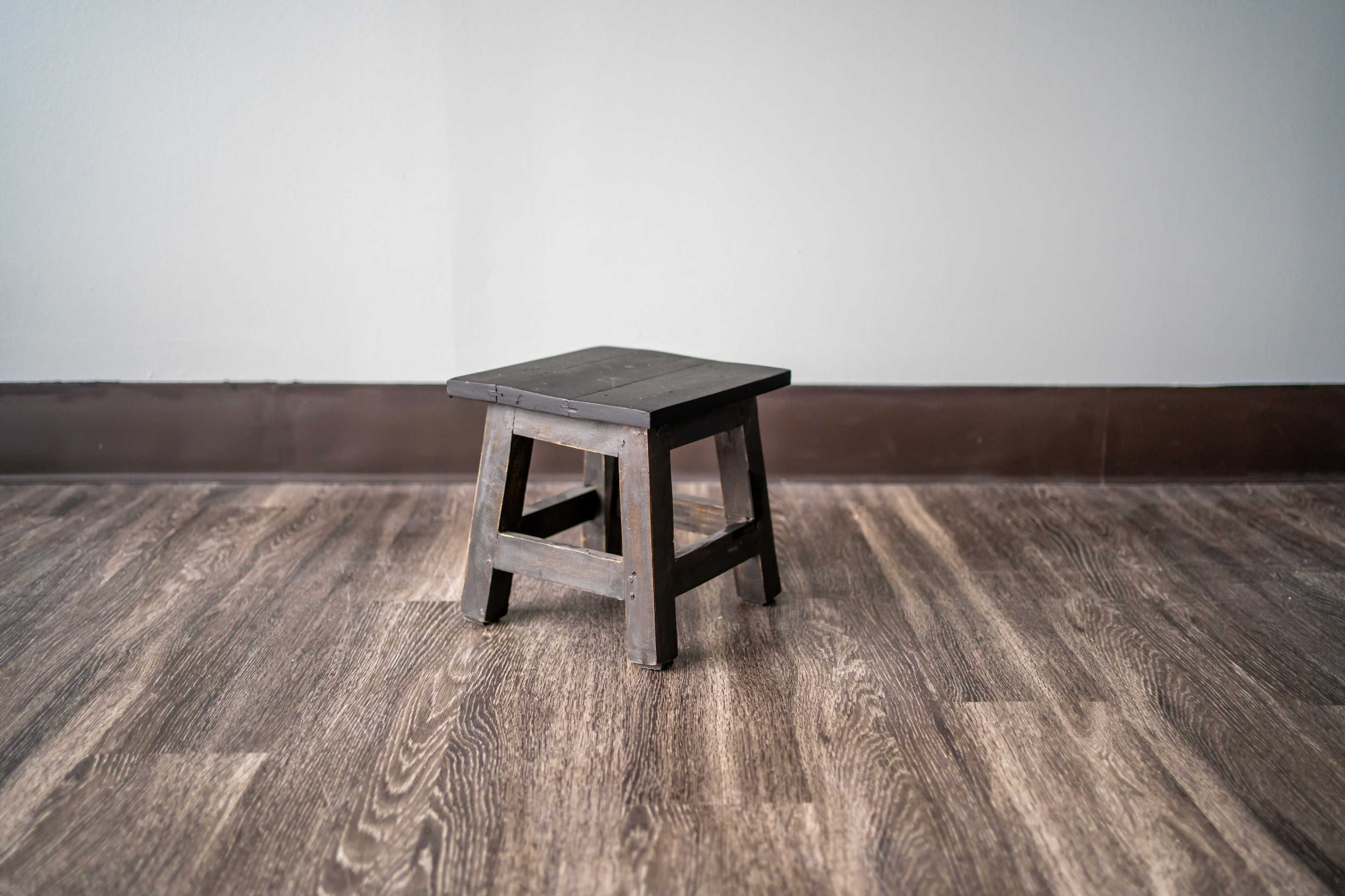 The image shows a small black wooden stool placed on a wooden floor against a gray wall.
