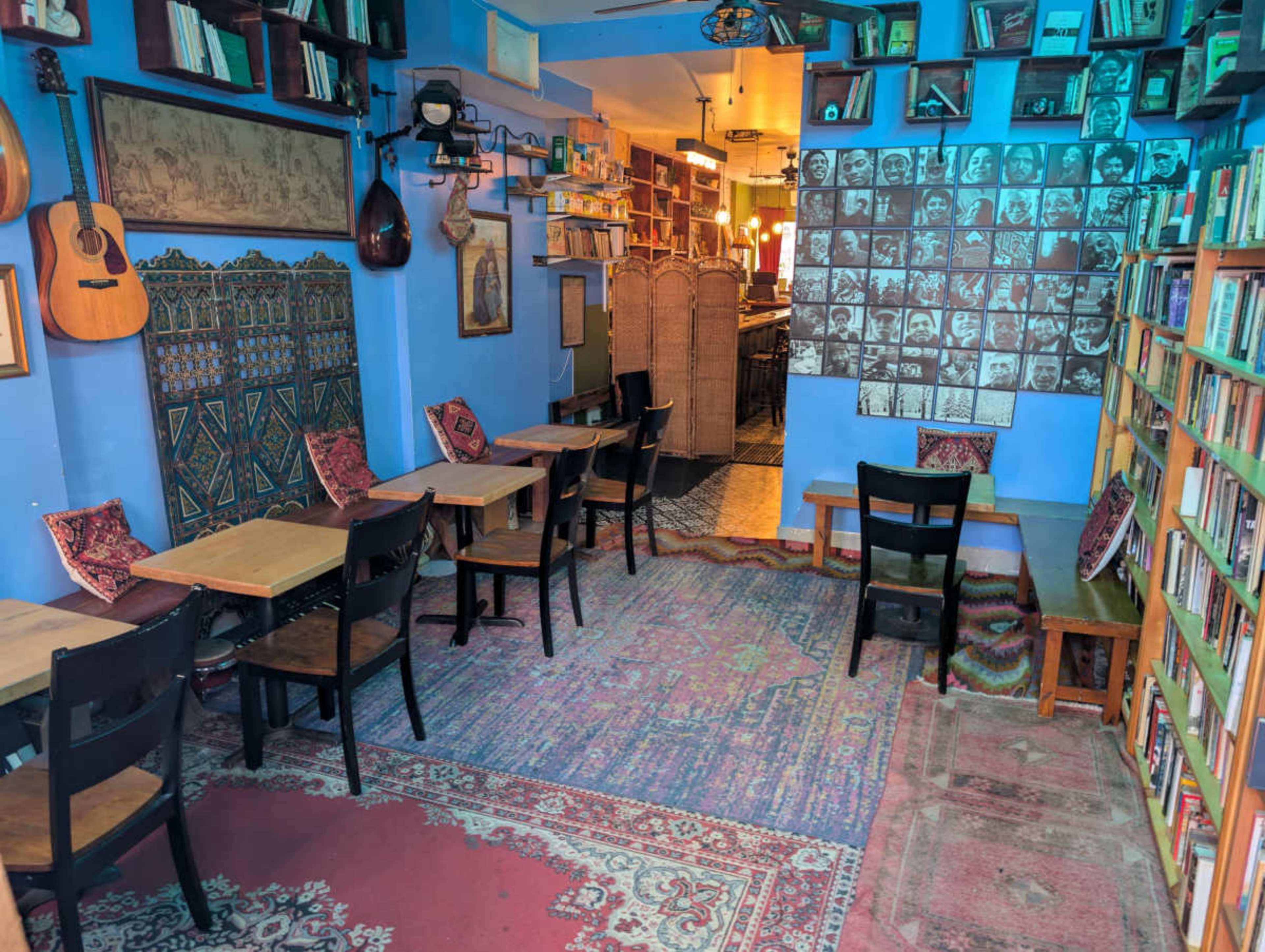 The image shows a cozy cafe with wooden tables and chairs arranged on patterned rugs, surrounded by shelves filled with books and adorned with musical instruments on the walls.