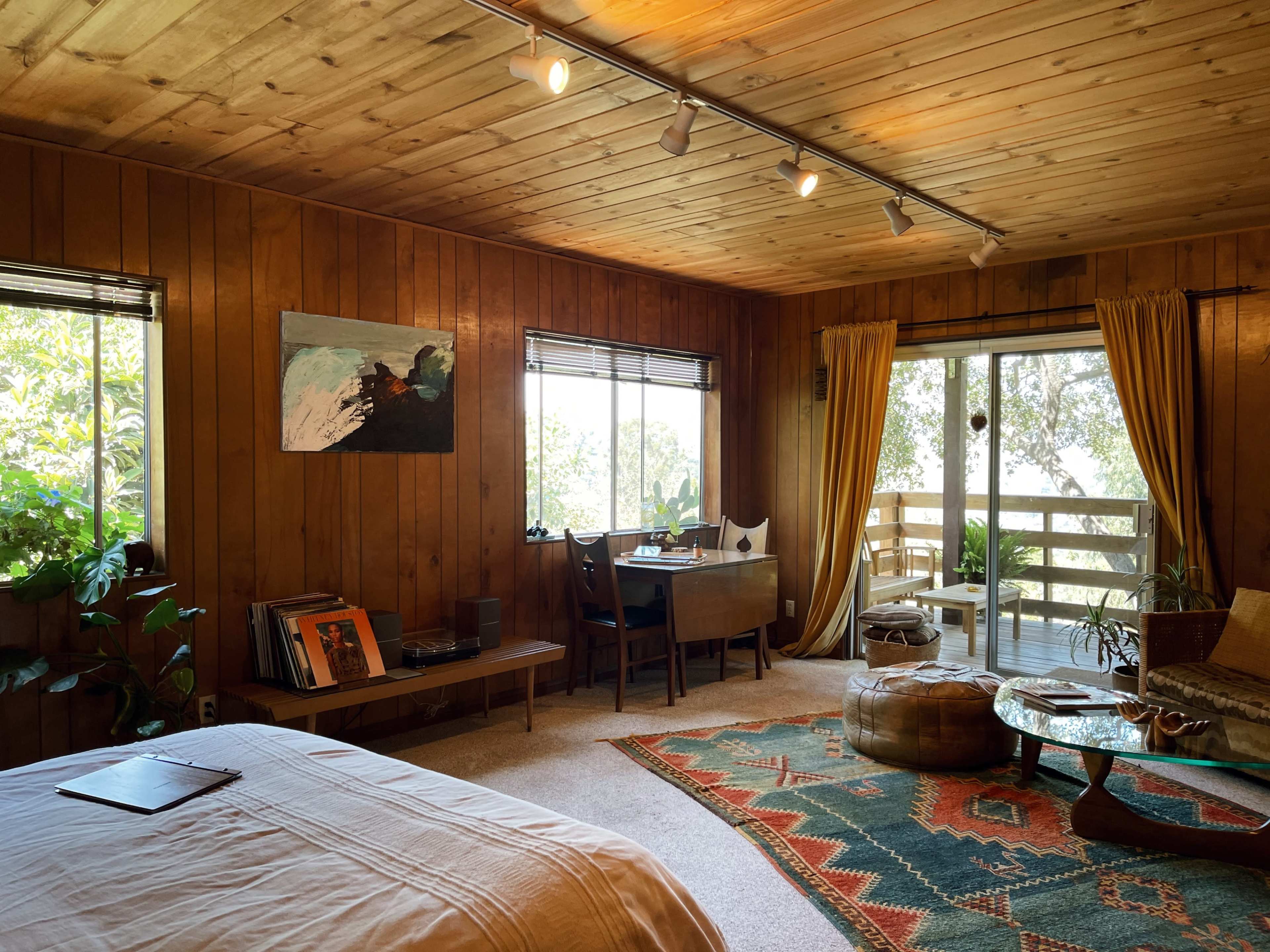 A rustic wooden bedroom featuring a bed, a desk with a chair, a seating area with a round table, and sliding glass doors leading to a balcony.