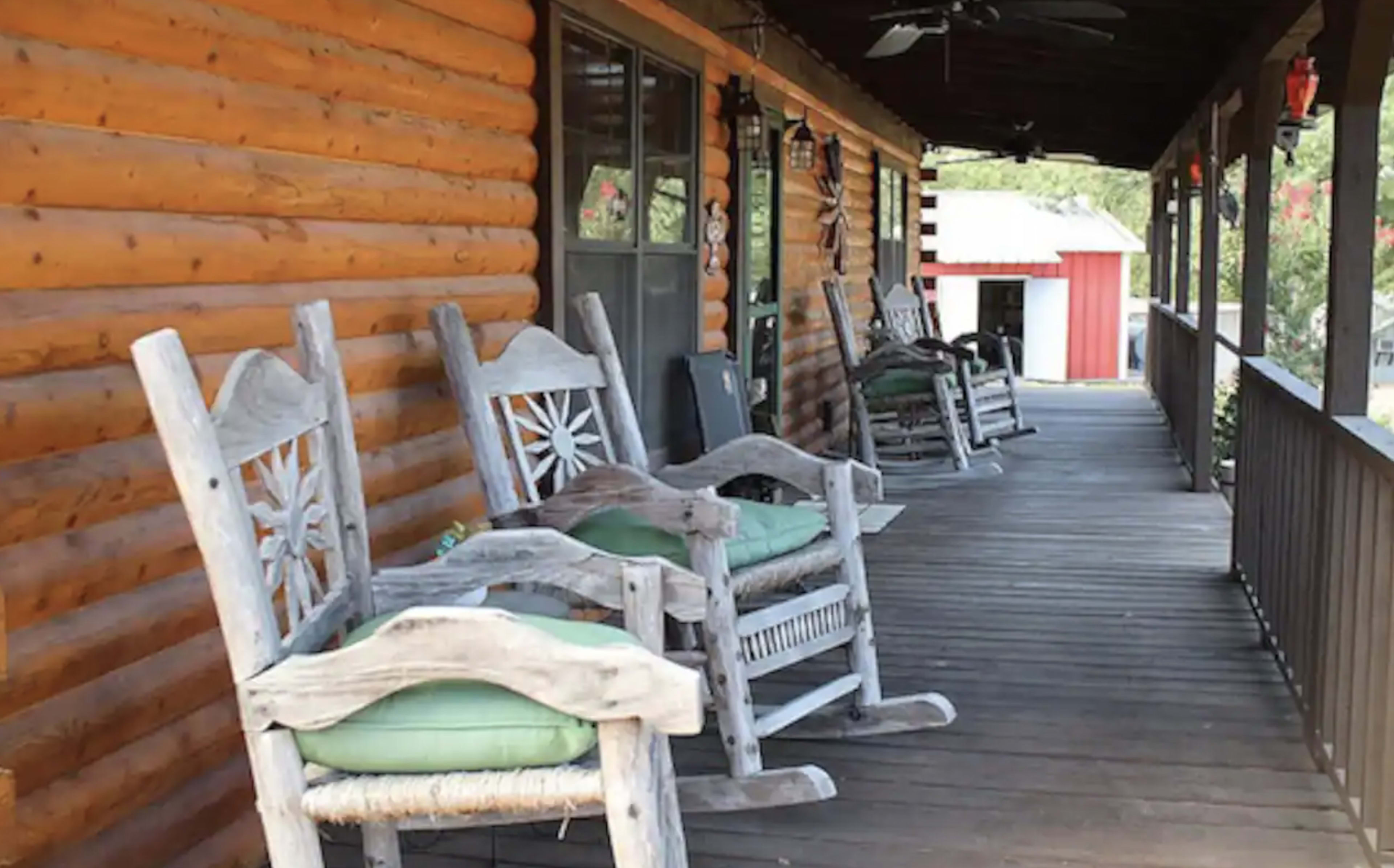 A wooden porch features several rocking chairs with green cushions, lined along a cabin-style log wall.