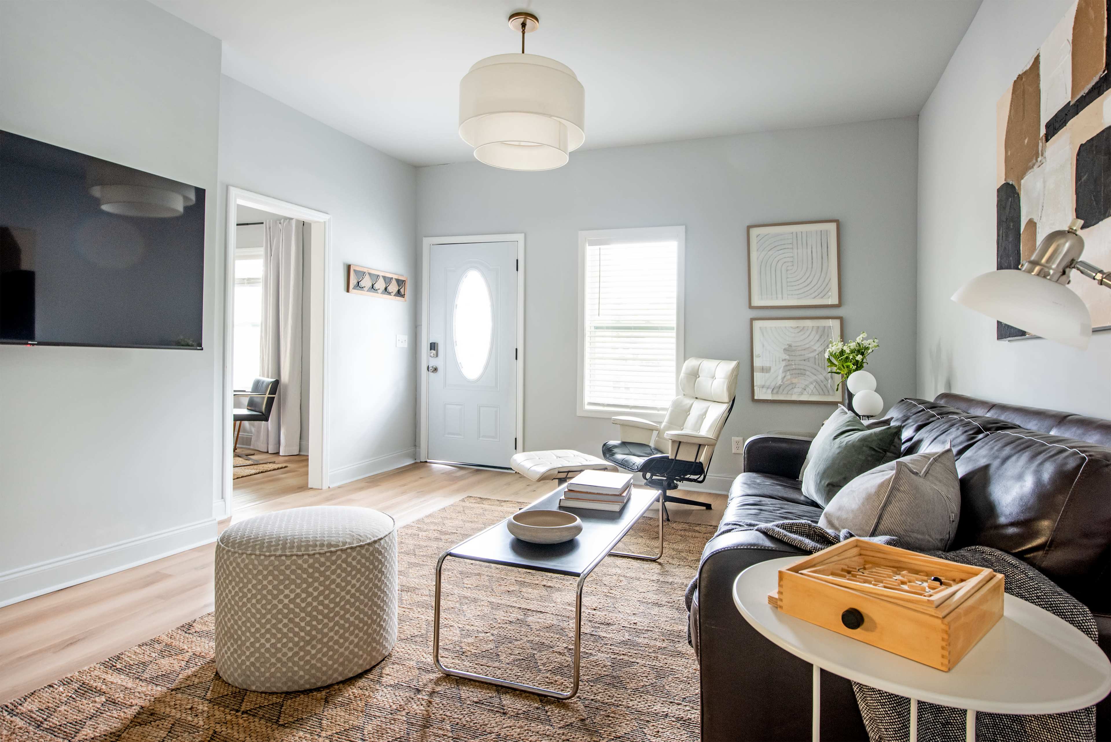 The image shows a modern living room featuring a gray sofa, a coffee table with books, a large television on the wall, and a round ottoman on a textured rug.