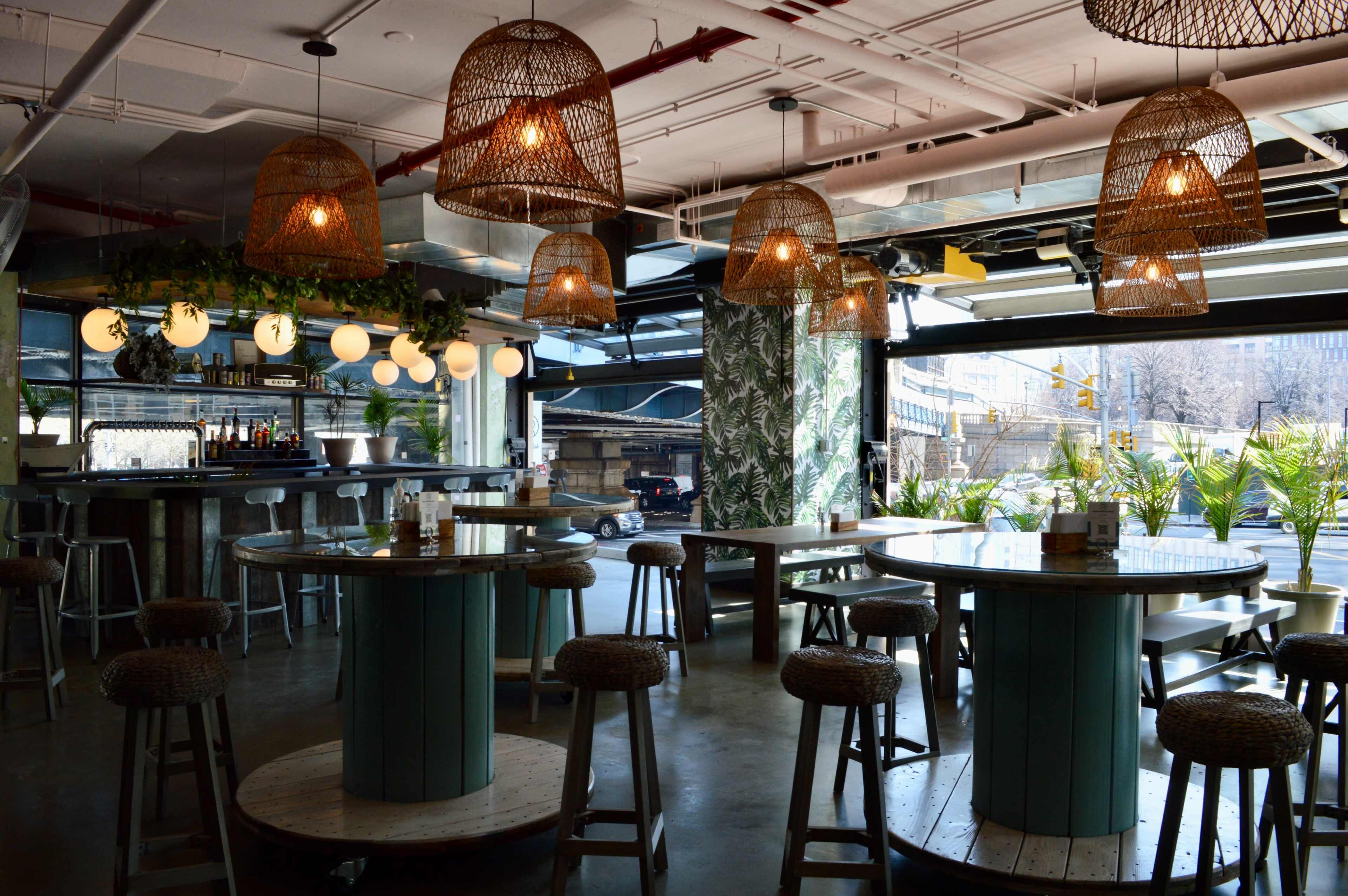 A modern restaurant interior featuring woven pendant lights, large wooden tables, and greenery by the windows.