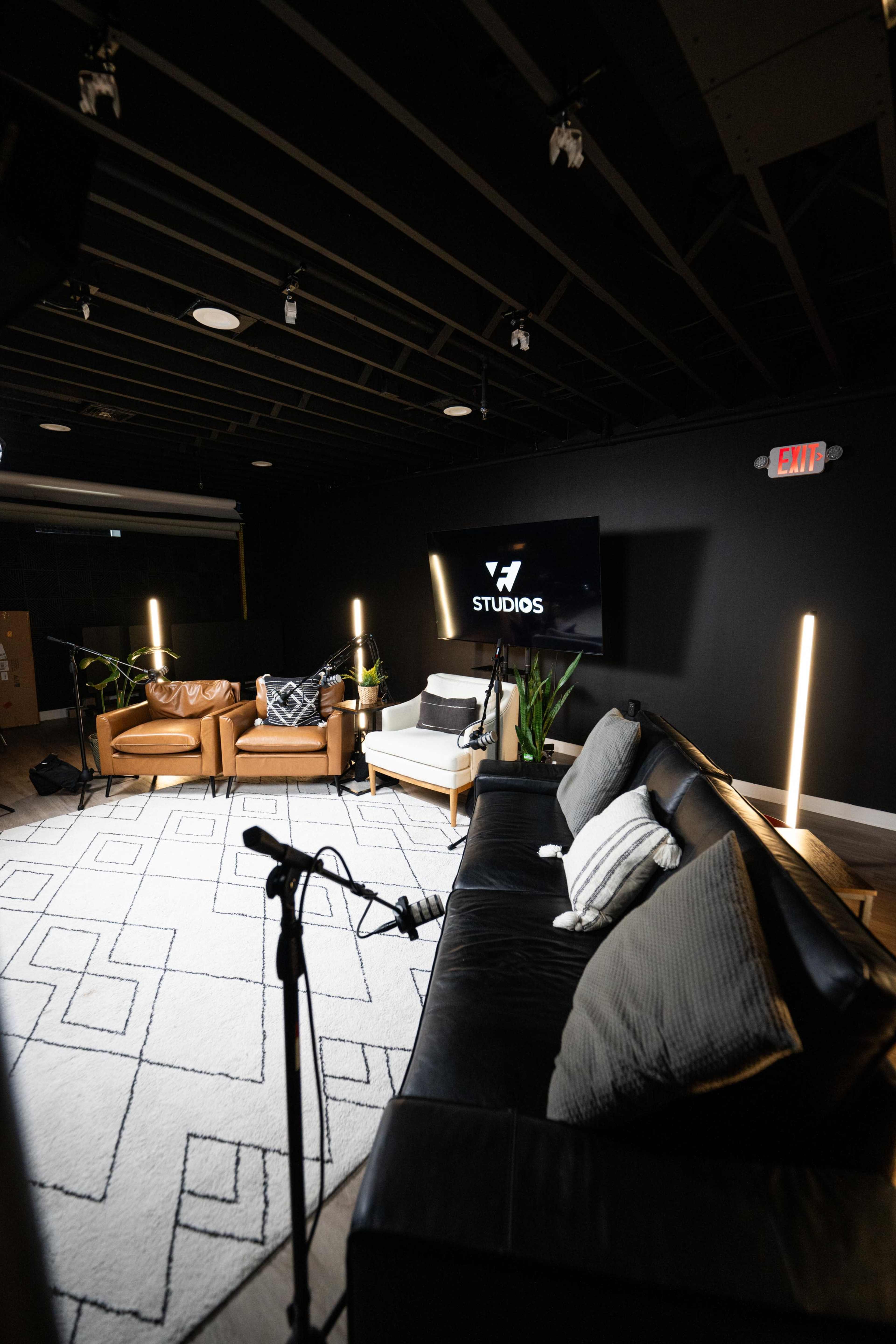 The image depicts a modern studio interior with a black wall, a large screen displaying the word "STUDIOS," and a combination of leather and fabric seating, all arranged around a geometric-patterned rug.