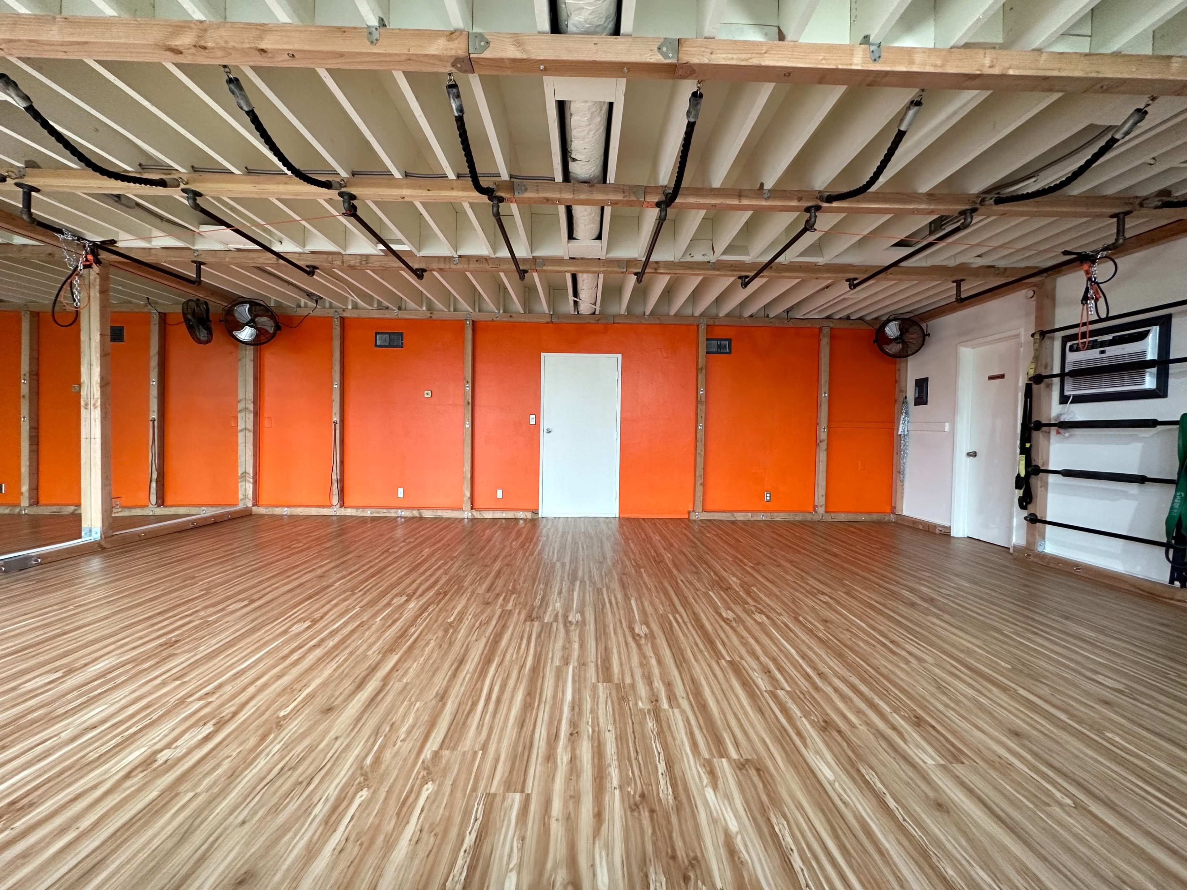 A spacious room with wooden flooring, orange-painted walls, mirrors on one side, and ceiling fans installed.