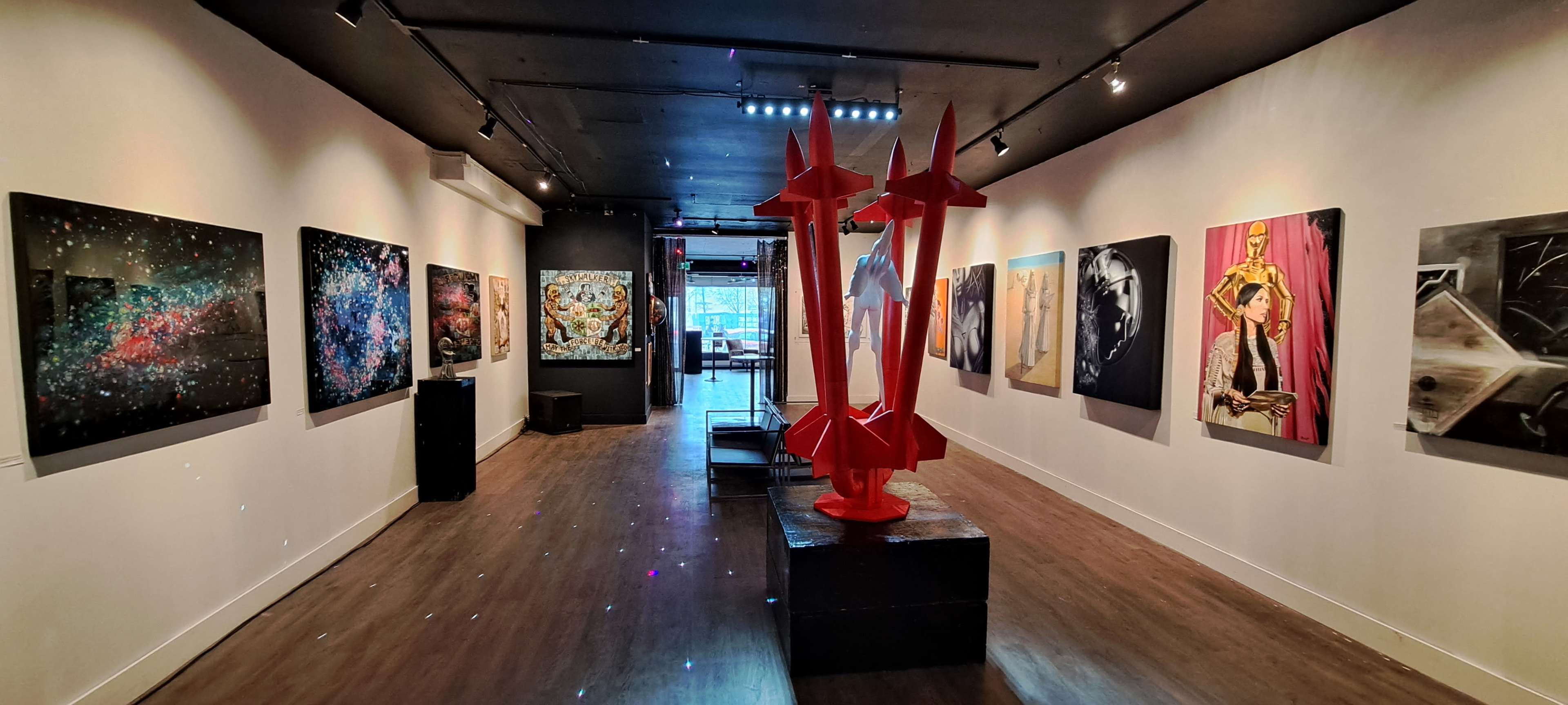 An art gallery interior featuring an array of paintings on the walls, with a prominent red sculpture in the center of the space.