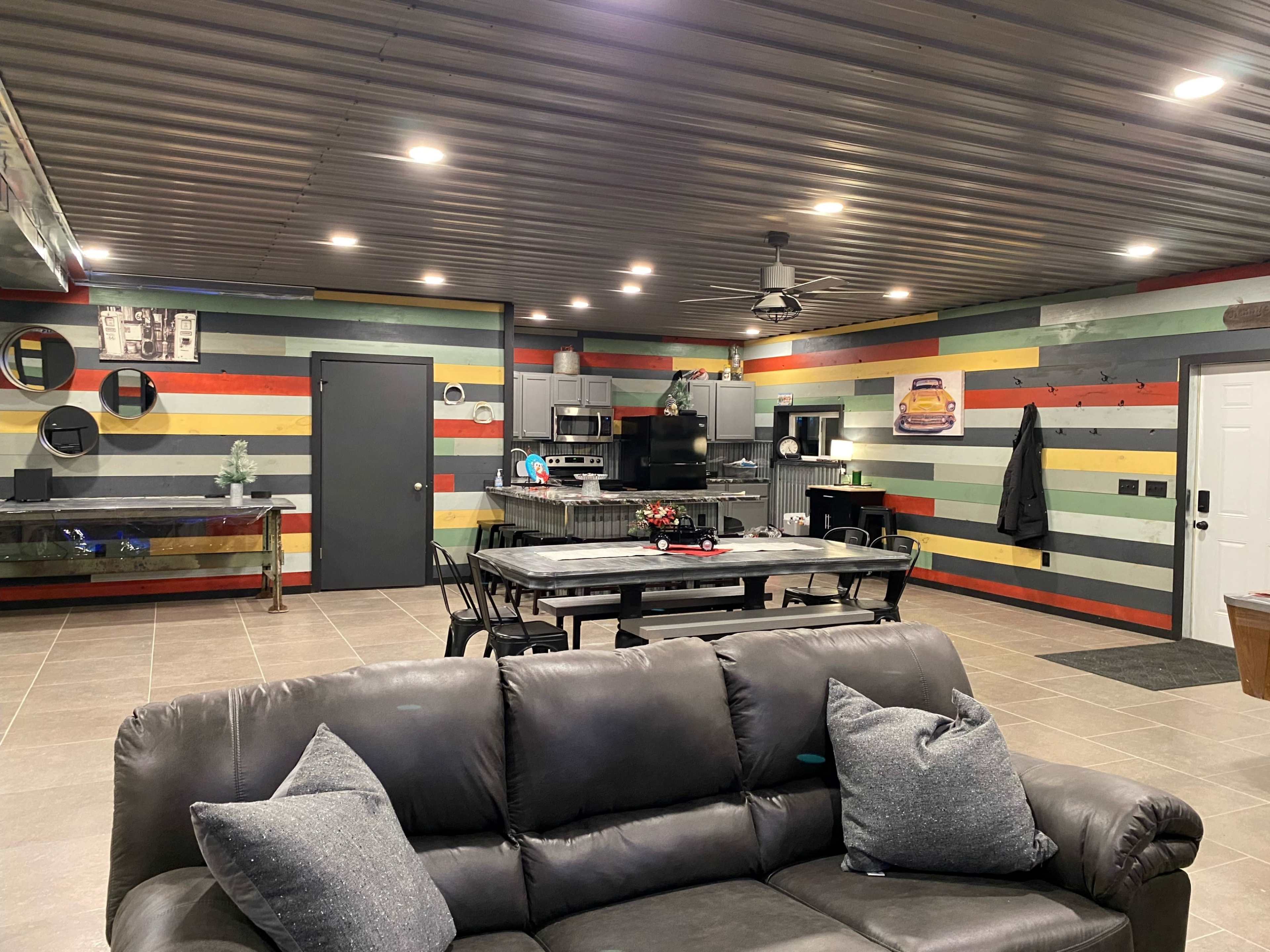 The image shows a modern, open-concept living space with colorful wooden wall stripes, a large dining table, and a kitchen area.