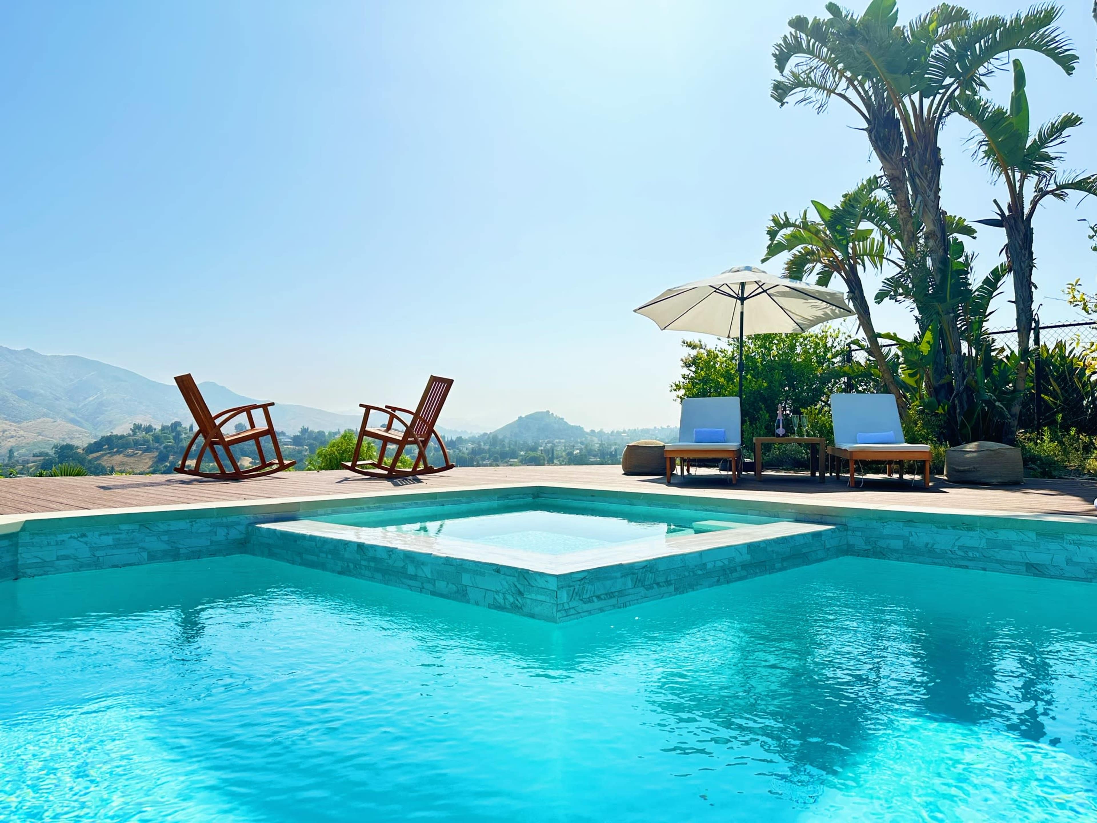 The scene features a sparkling swimming pool surrounded by wooden lounge chairs and a sun umbrella, set against a backdrop of hills and clear blue skies.
