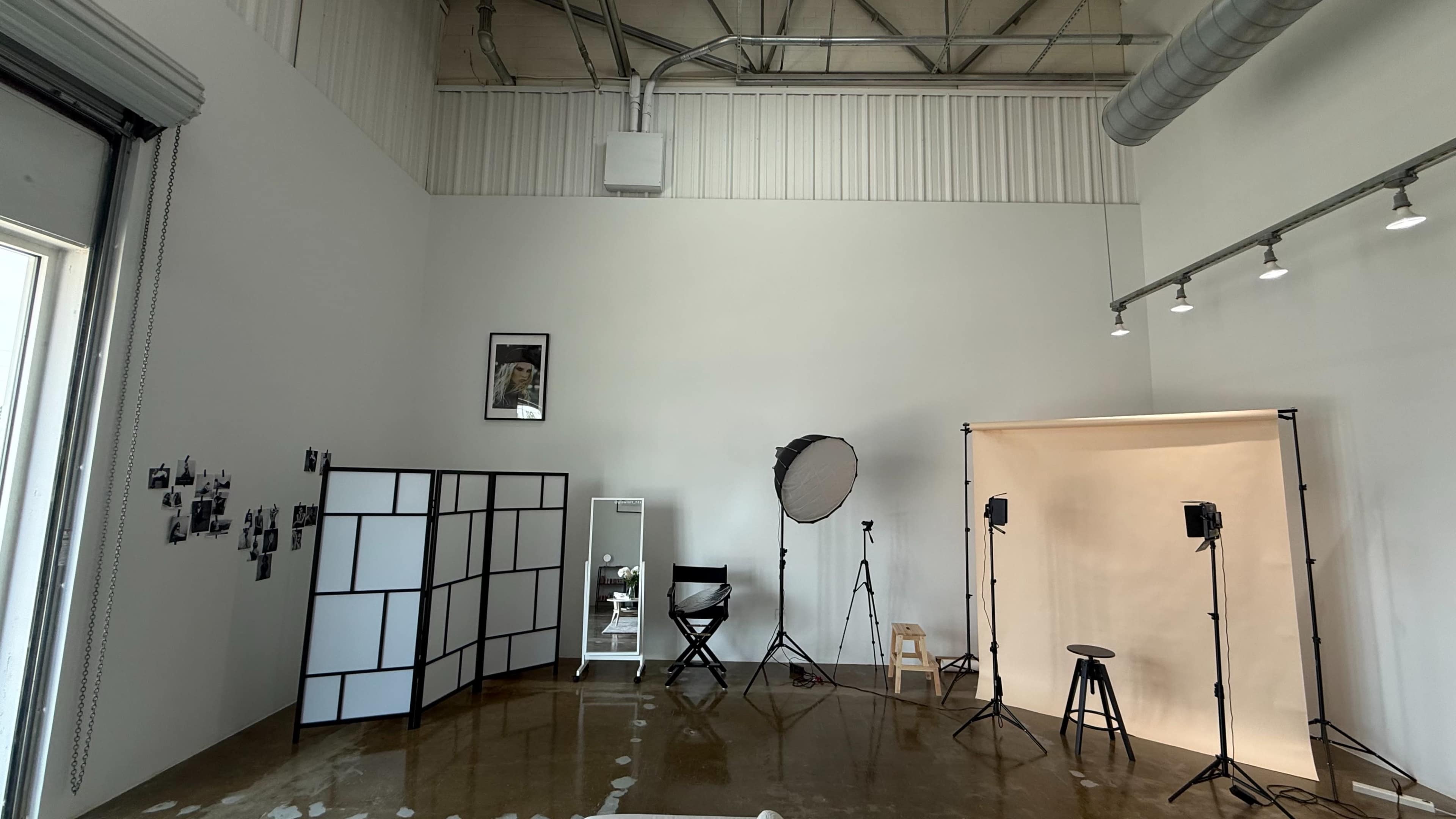 The image shows a spacious photo studio with a light-colored backdrop, various lighting equipment, a director's chair, and a partition displaying black-and-white photographs.